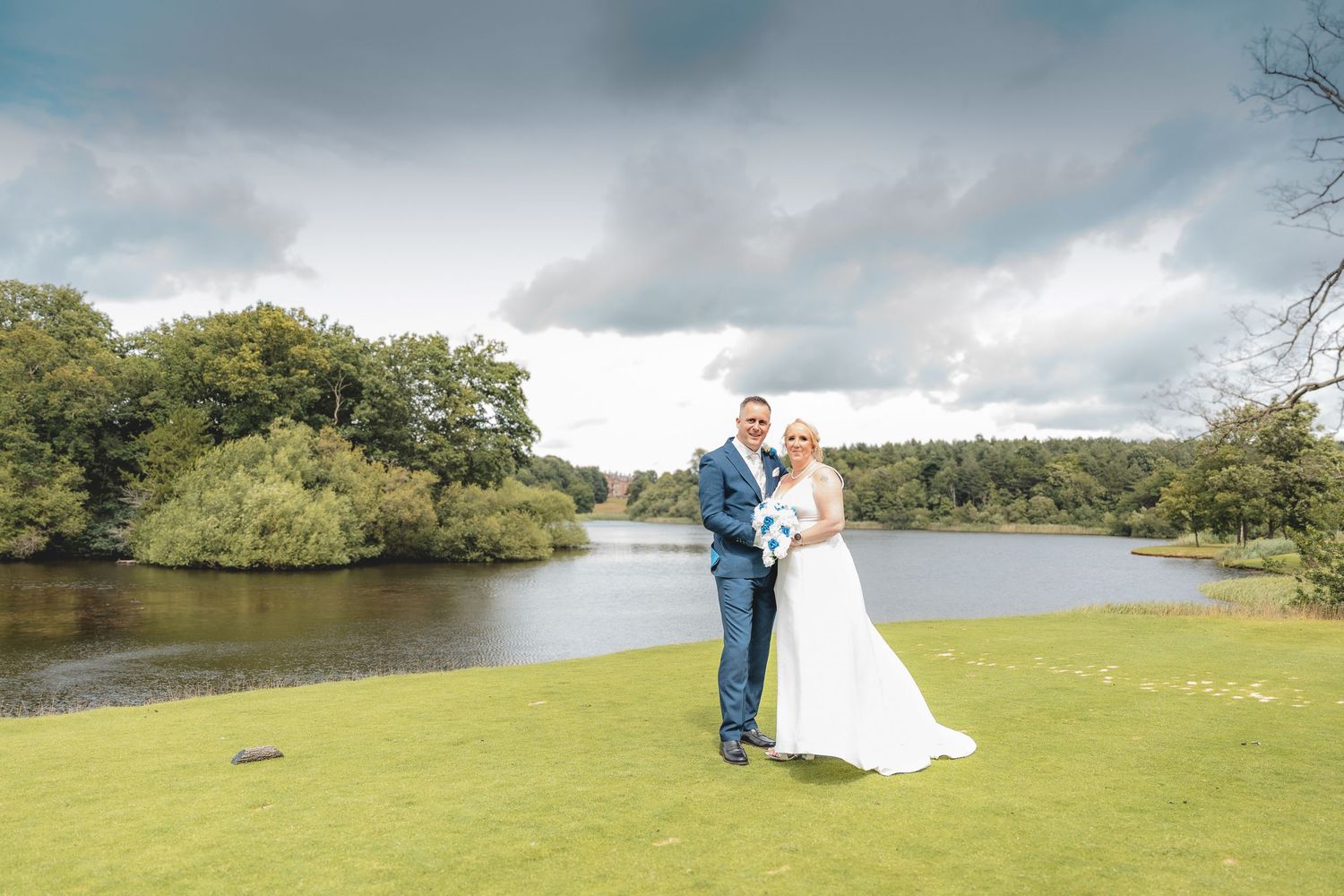 A couple in wedding attire poses together on a grassy lawn beside a serene lake under dramatic cloudy skies.