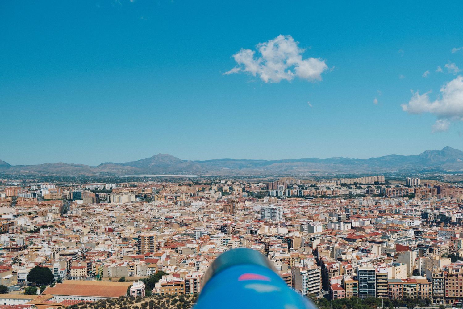 Panoramic cityscape of Alicante with mountains in the background and a blue telescope in the foreground.