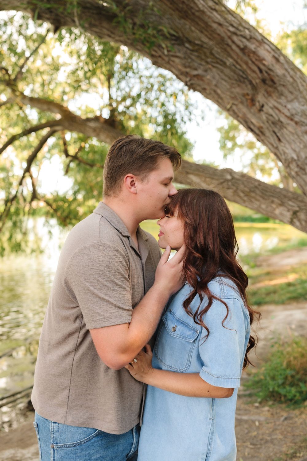 A romantic couple shares tender moments in a natural outdoor setting under a large tree.