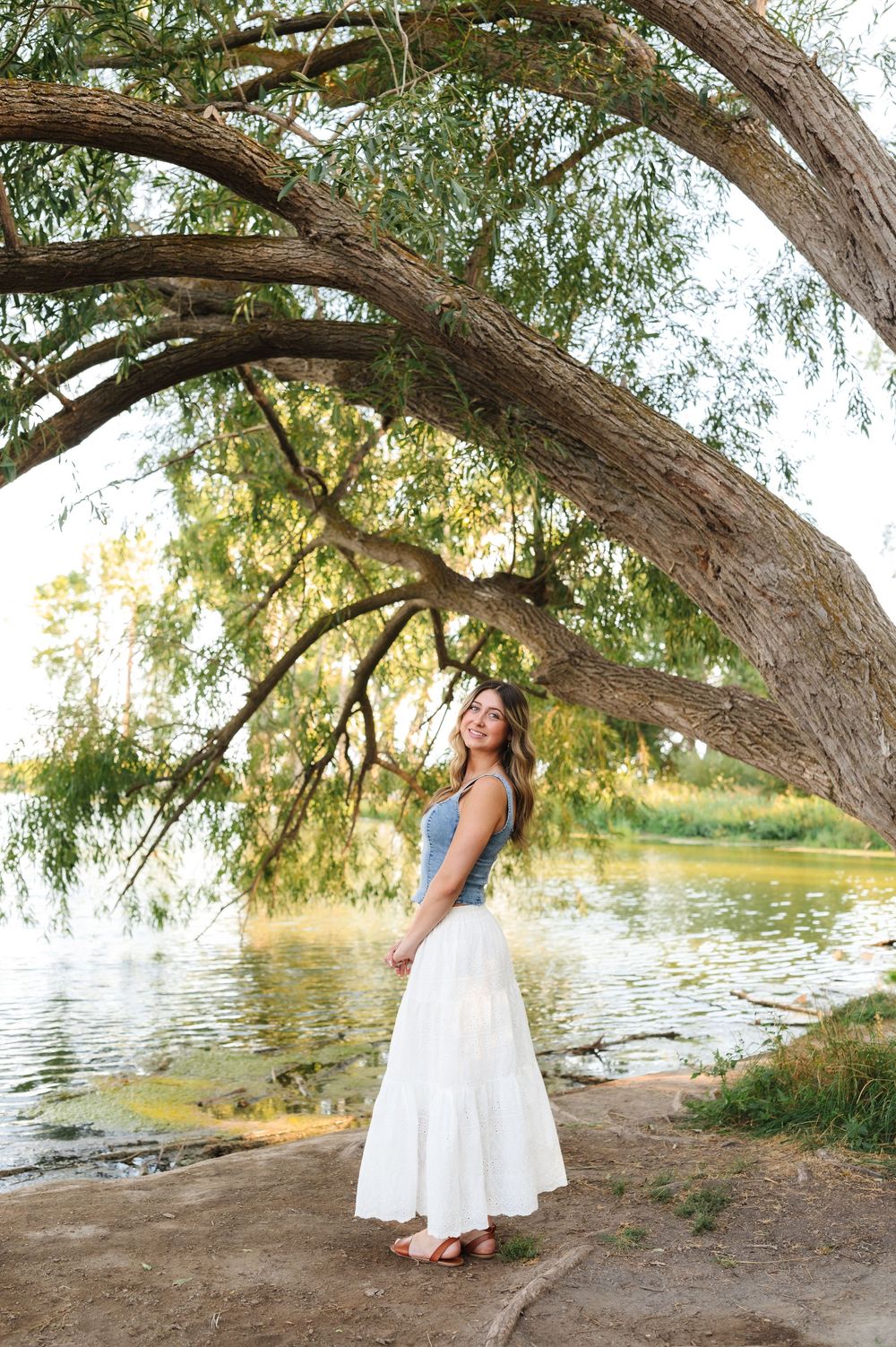 A white sundress under a willow tree by a serene lake creates a dreamy summer photoshoot scene.
