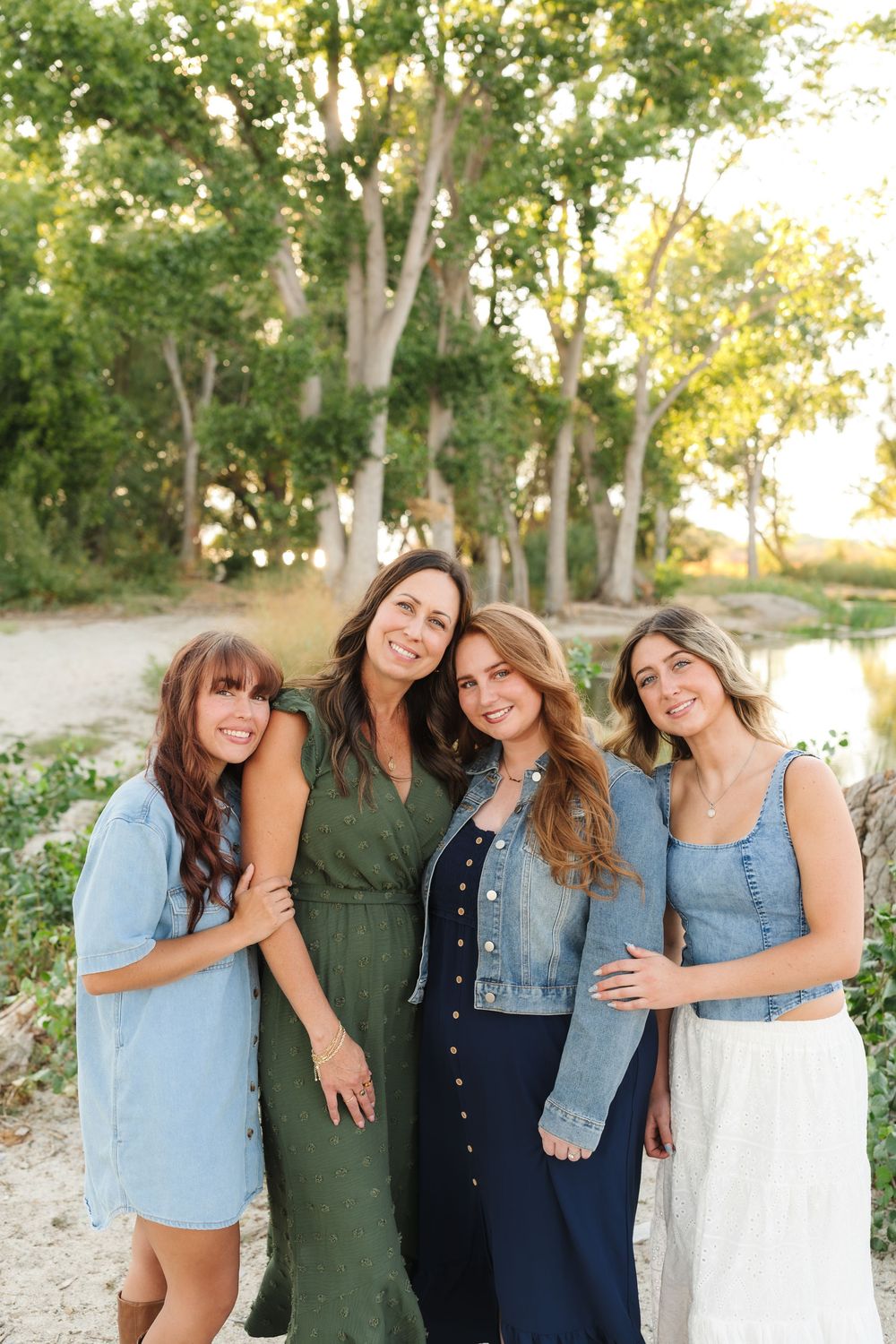 Four friends in casual denim and olive outfits pose together outdoors against a natural wooded background at sunset.