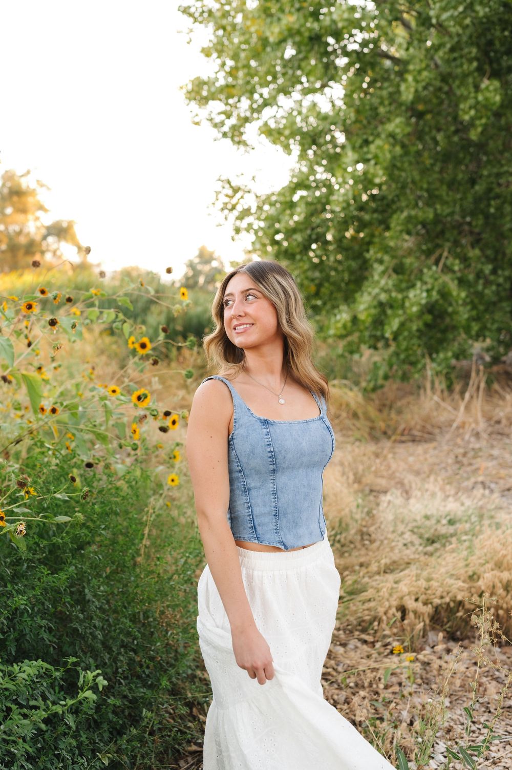 A person in a denim crop top and white skirt stands in a sunny meadow with yellow wildflowers and green trees.