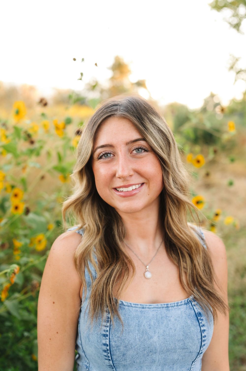 A smiling woman in a denim top poses in front of a field of sunflowers during golden hour.