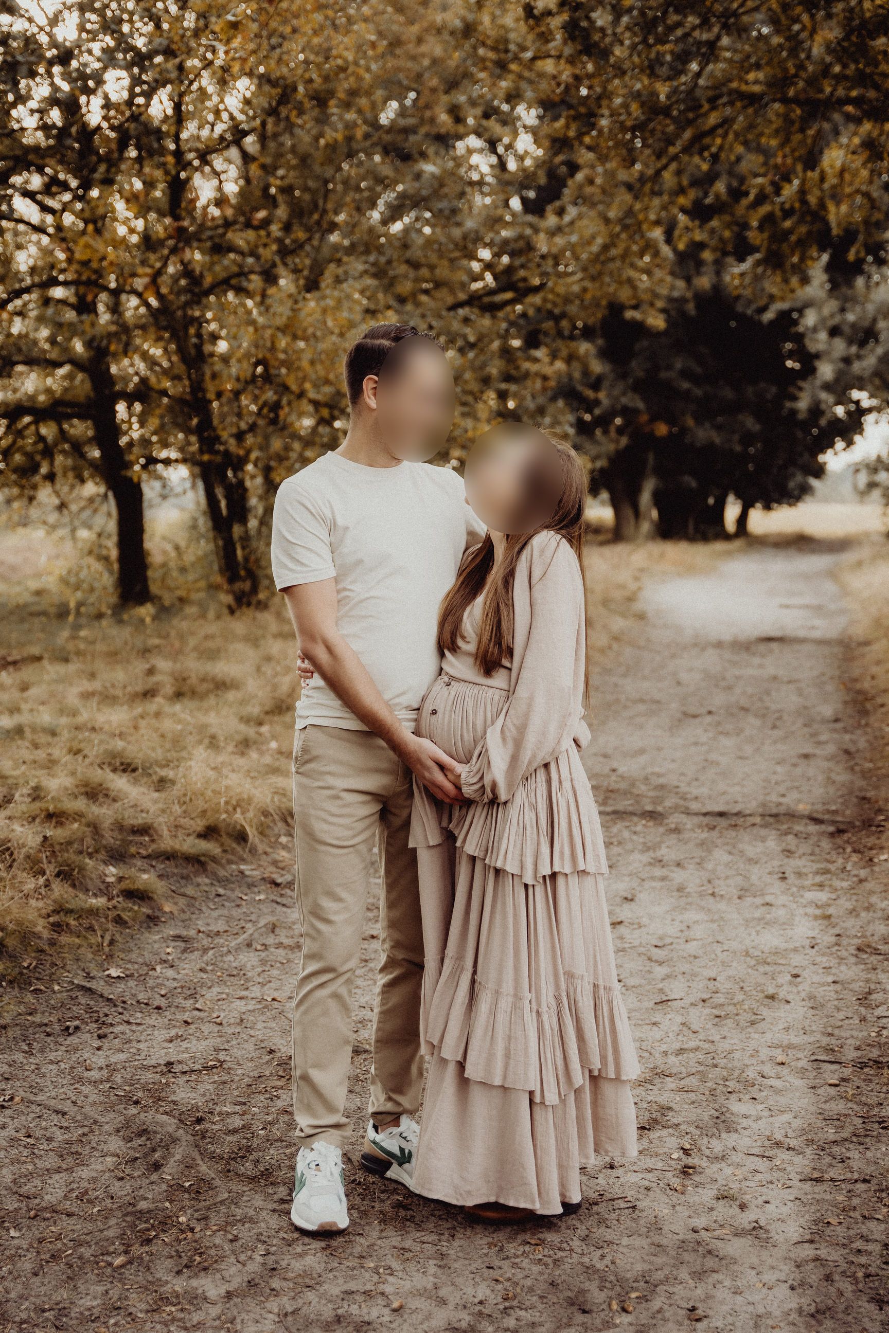 Two figures in neutral tones embrace on a dirt path surrounded by autumn trees at golden hour.