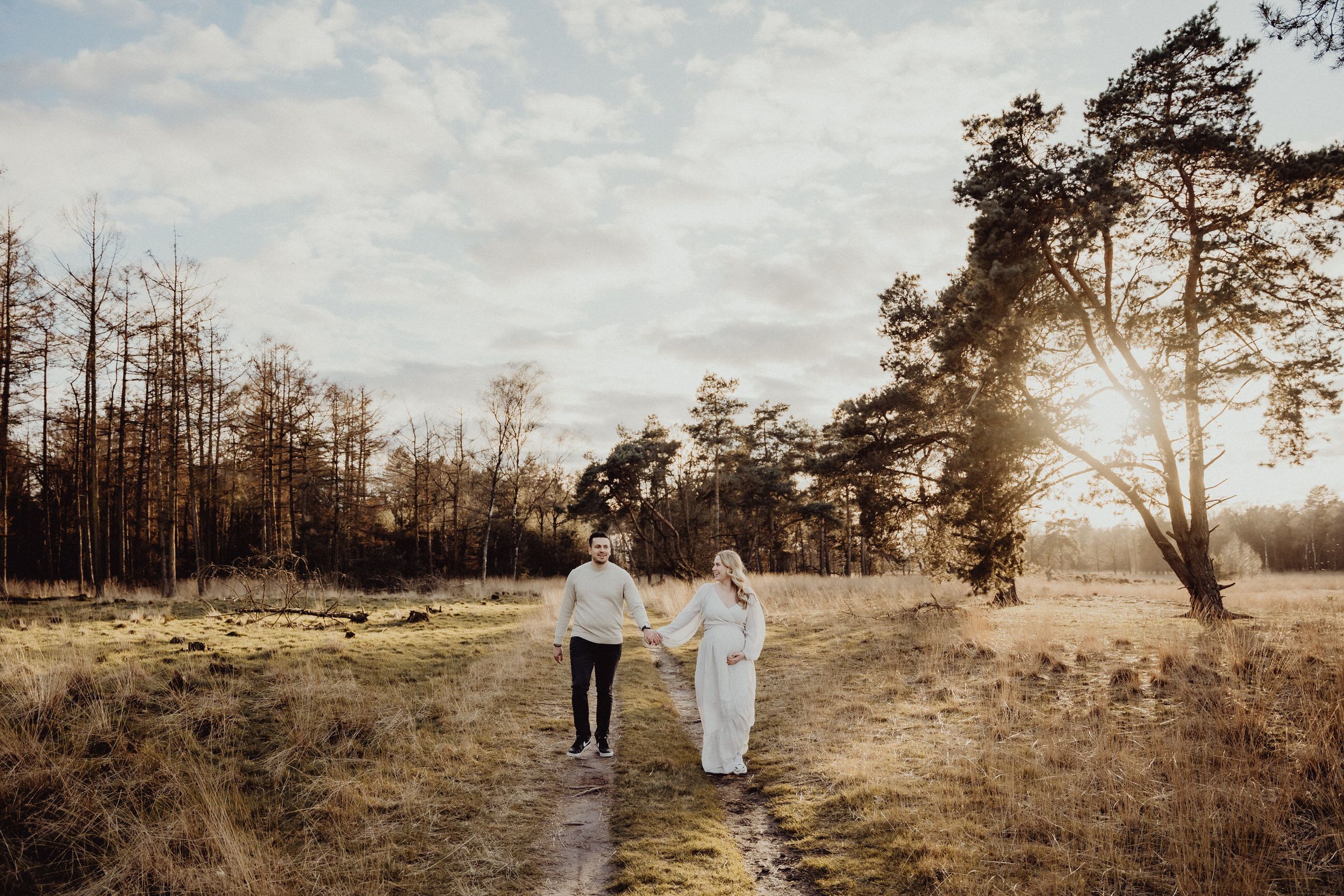 Two figures walk along a nature path with sunlight filtering through pine trees.