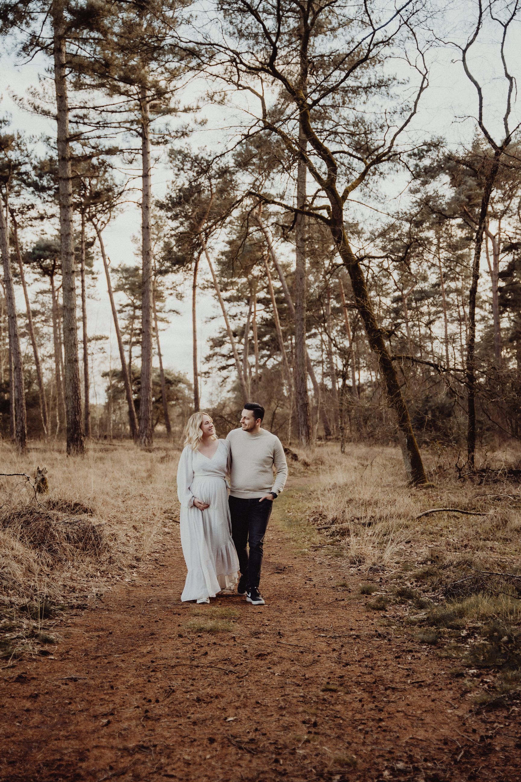A couple walks hand in hand down a forested path surrounded by tall pine trees.
