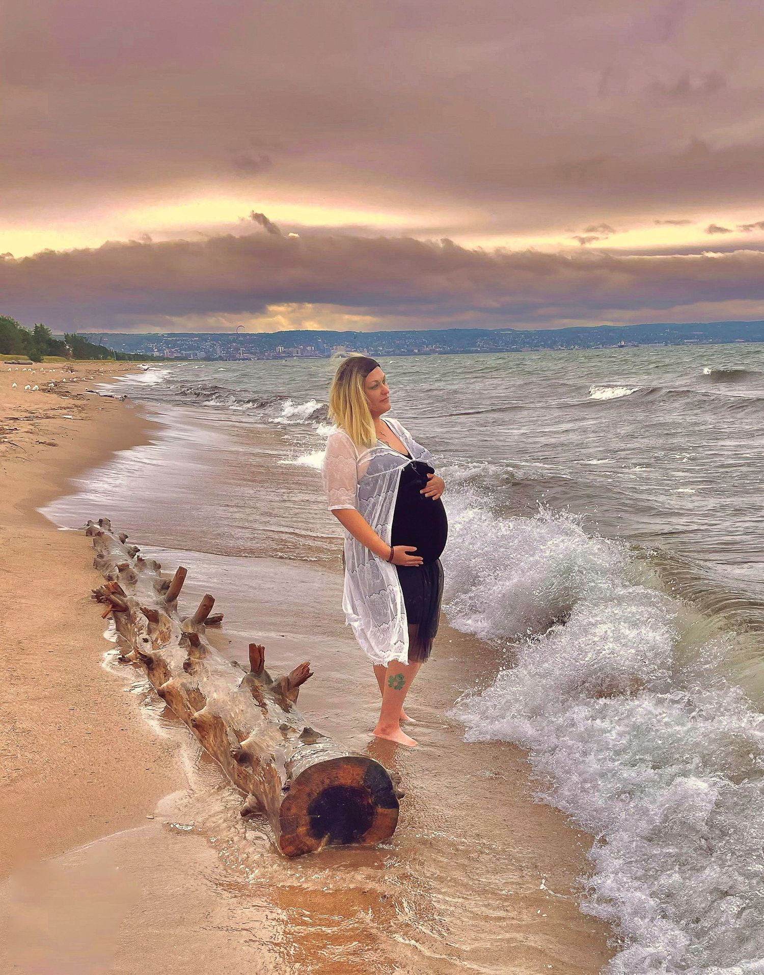 A dreamy beach themed maternity shoot at sunset with waves crashing against driftwood on the shore.