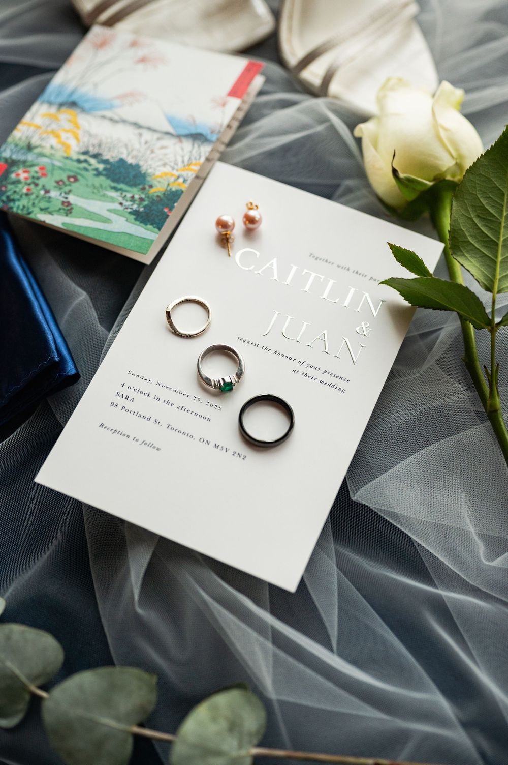 Wedding rings and invitation cards displayed on gray fabric with delicate white roses and greenery.