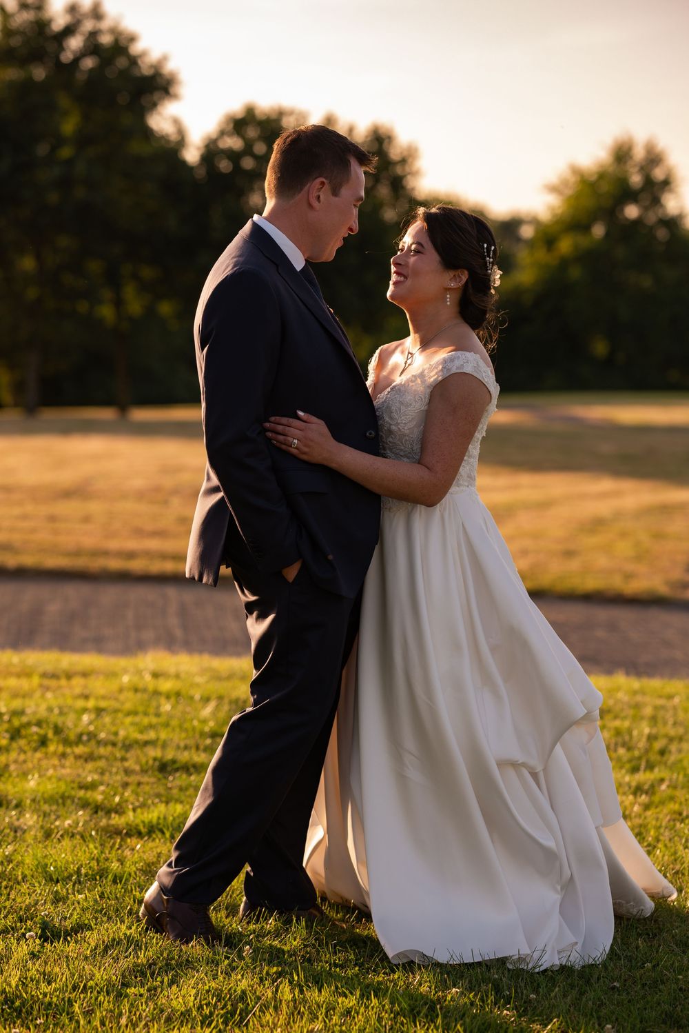 A couple in wedding attire embraces on a grassy field during golden hour sunset.
