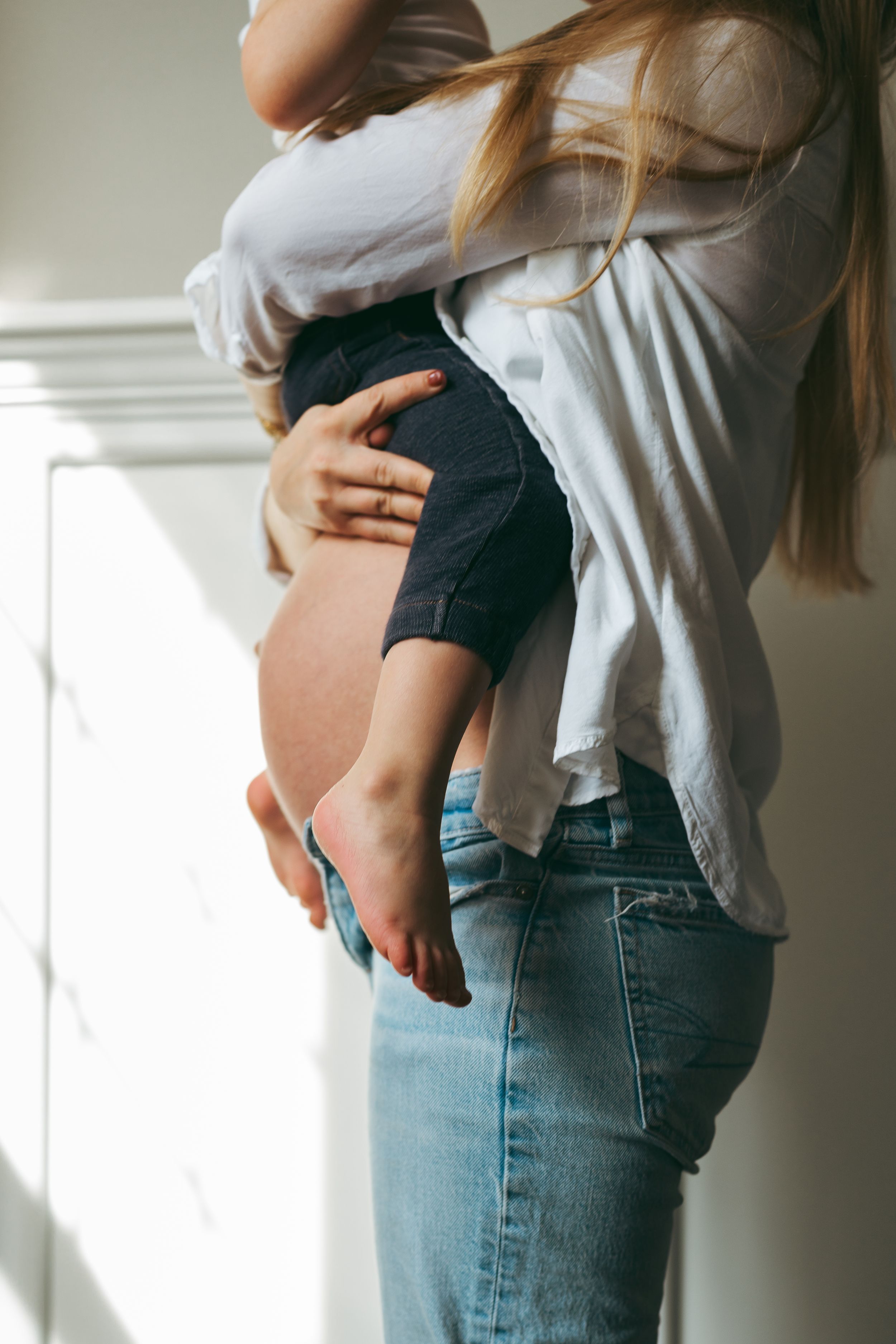 Young child being held and hugged while wearing a white shirt, standing in natural window light.