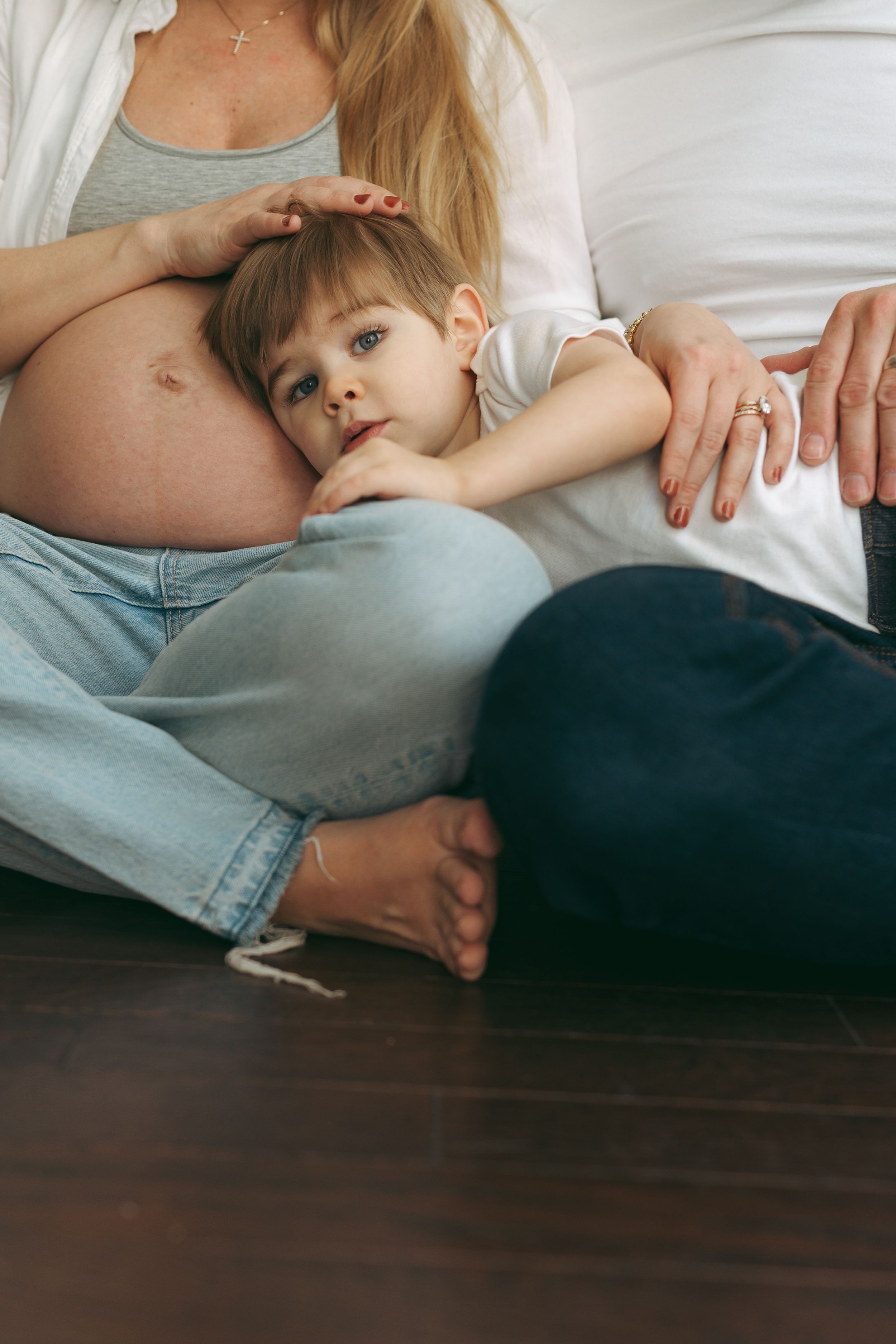 A young child rests their head on a pregnant belly while sitting on hardwood floor.