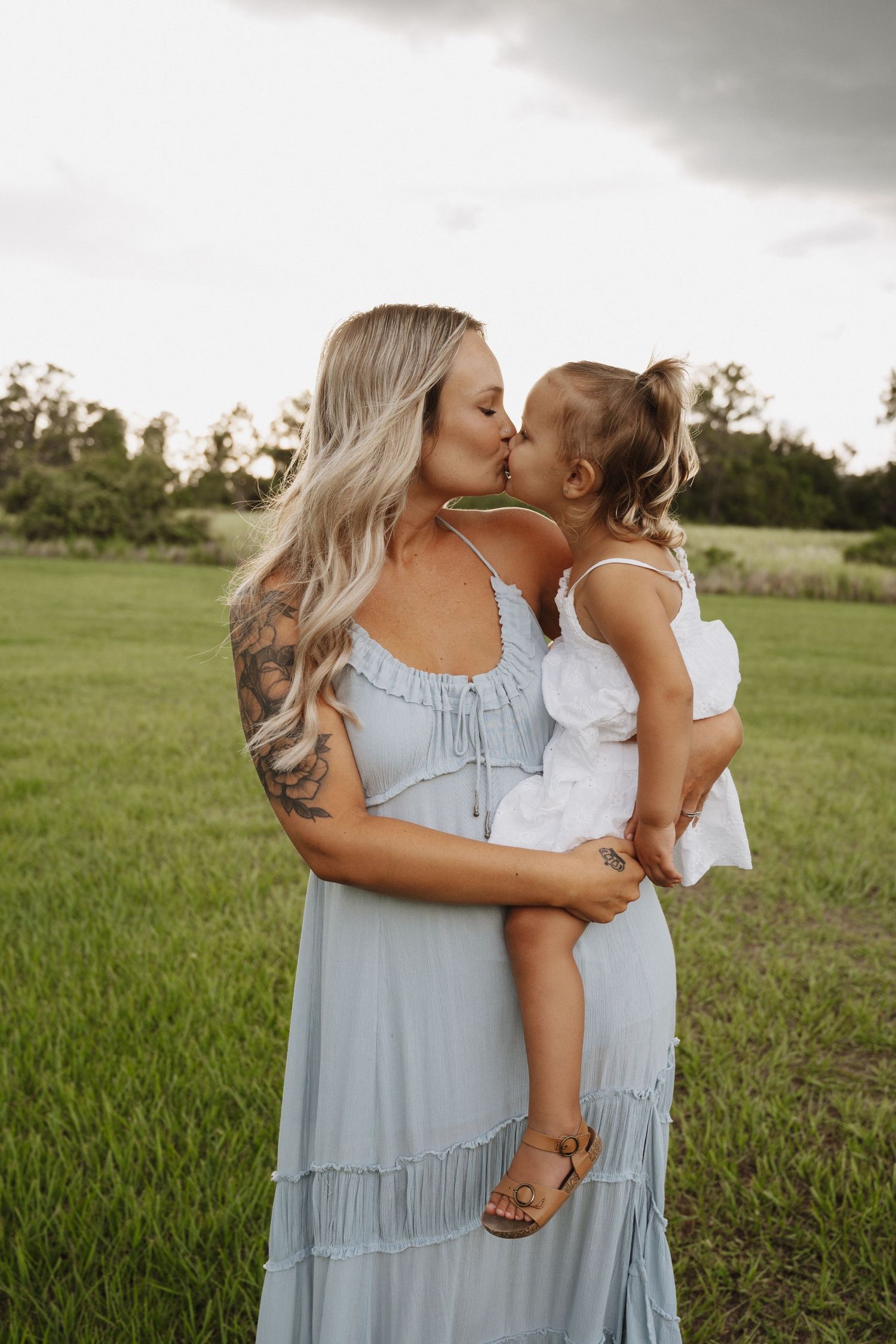 Two figures in light blue dresses embrace tenderly in a sunlit green field during golden hour.