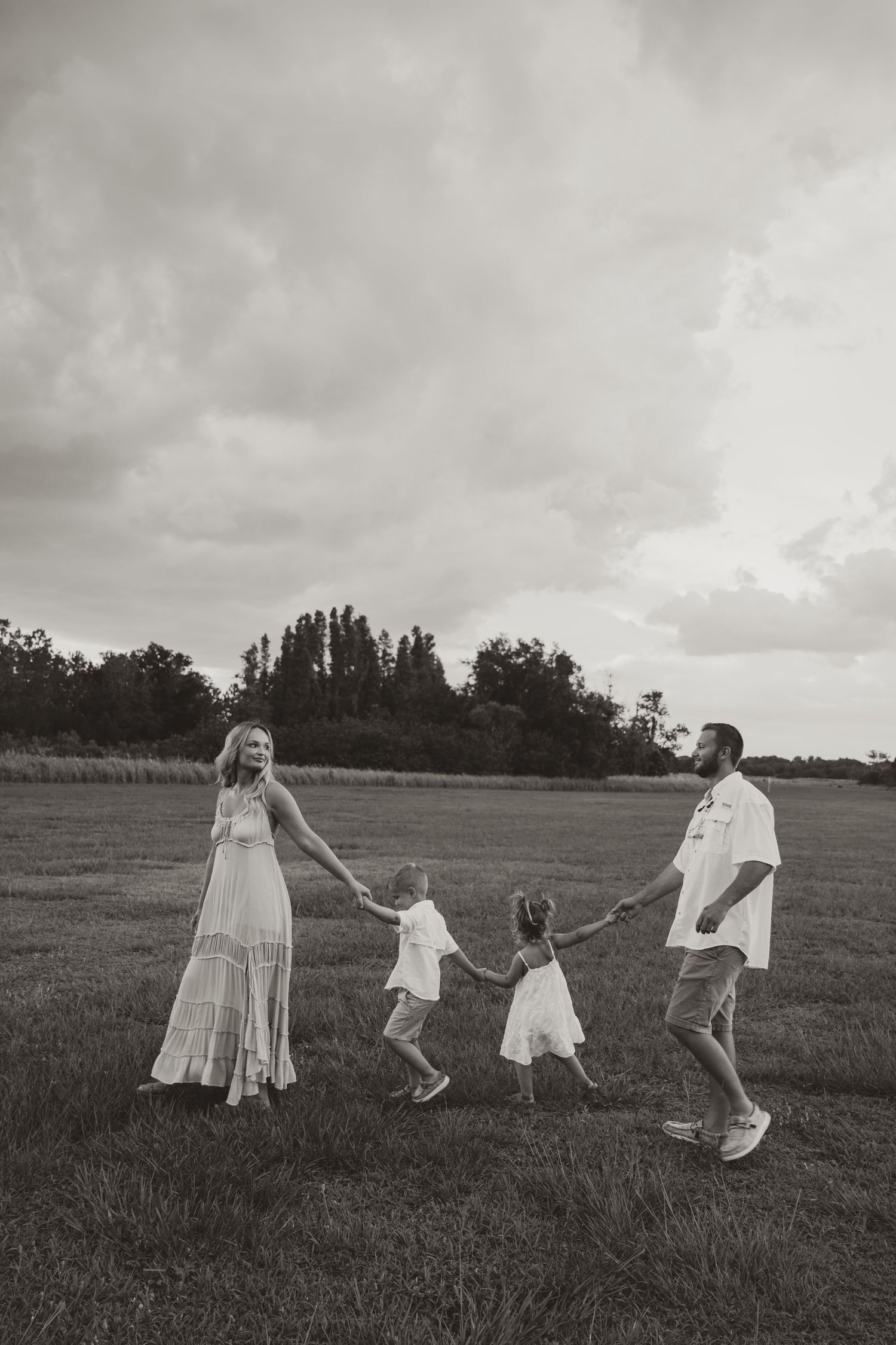 A family holds hands while joyfully skipping through a grassy field under cloudy skies in black and white photography.