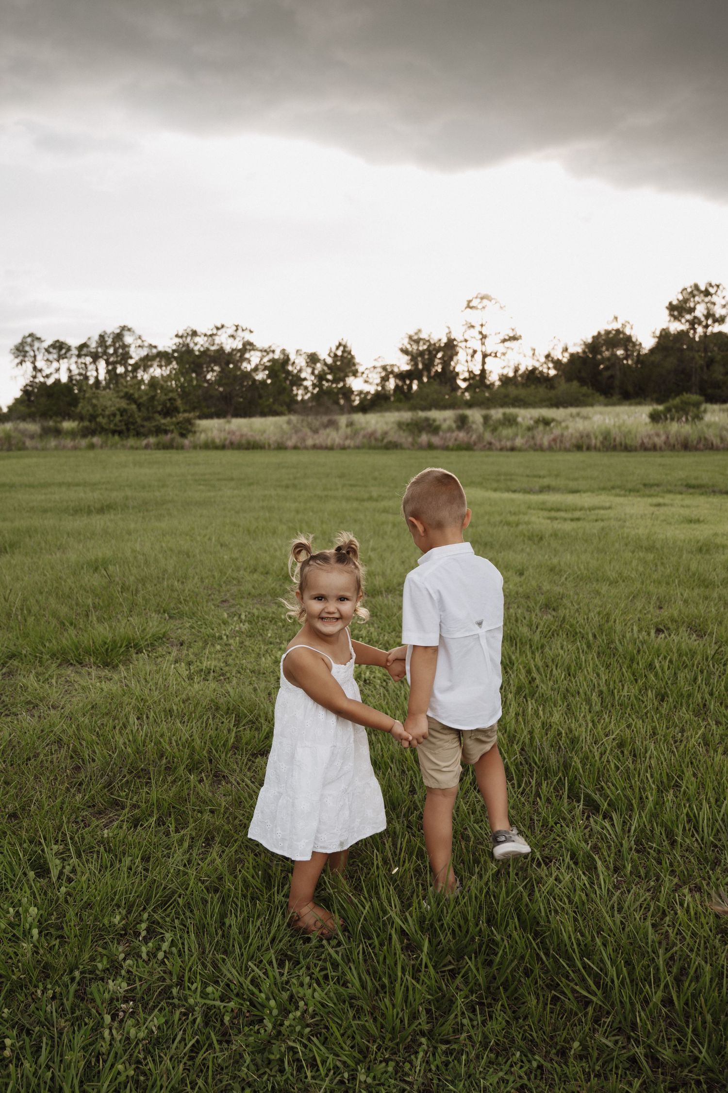 Two small children hold hands while walking together through a green grassy field under moody storm clouds.