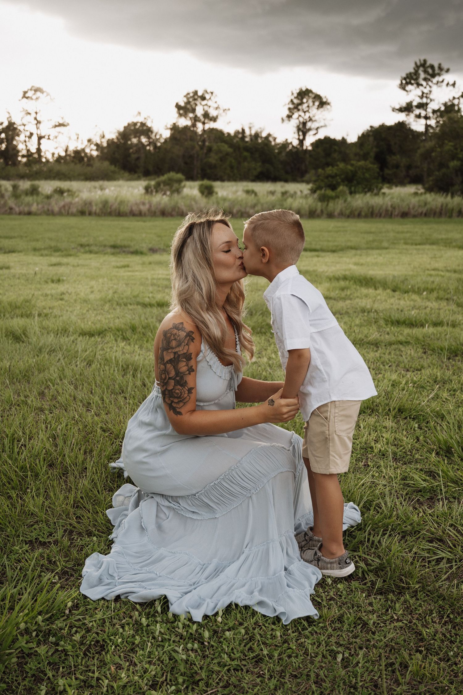 A tender moment captured between parent and child in a grassy field at dusk with pine trees in the background.