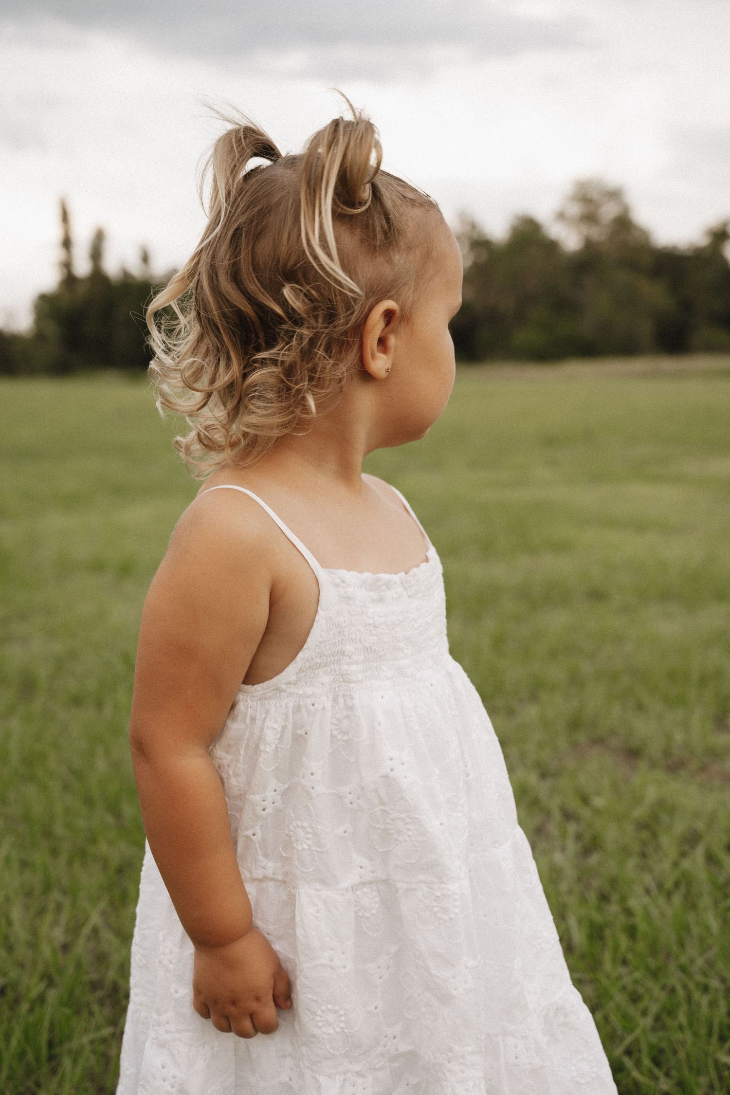Small child in a white tiered summer dress stands in a grassy field on a cloudy day looking to the side.