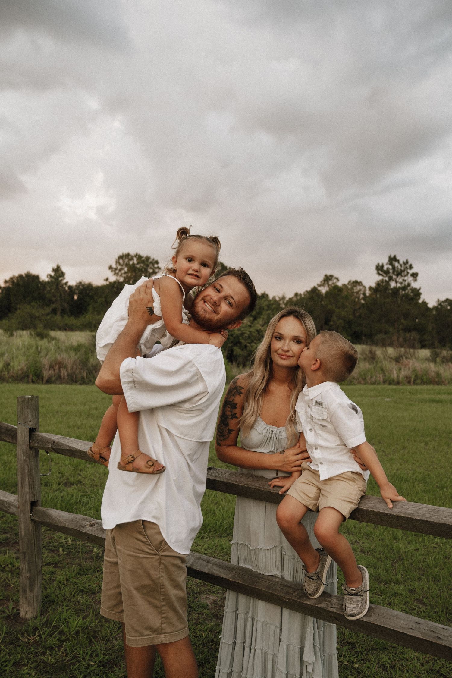 Family shares joyful candid moments together while posing along a rustic wooden fence in a green meadow under moody skies.