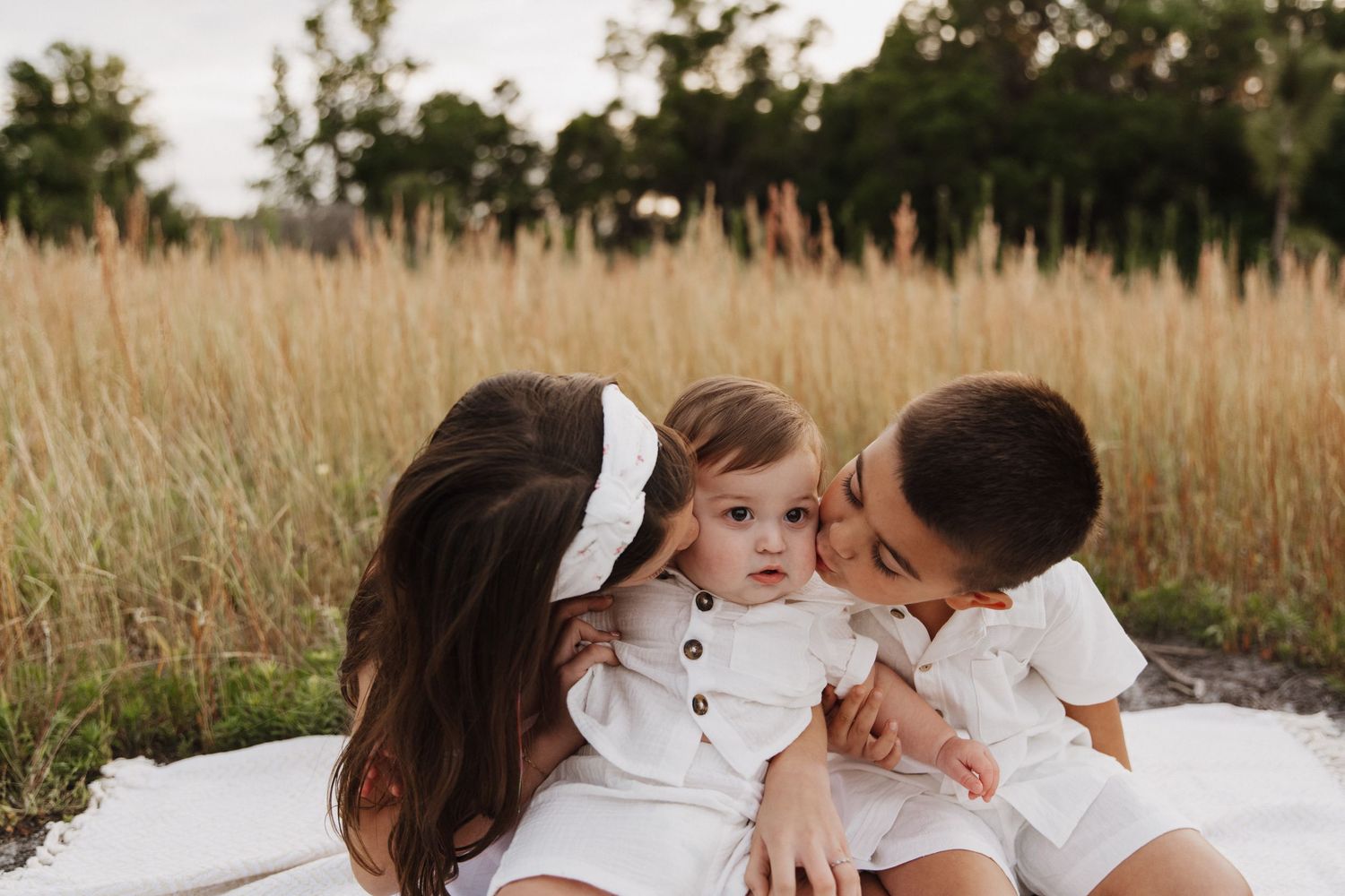 Three young children in white outfits sitting on a blanket in a golden wheat field at sunset.