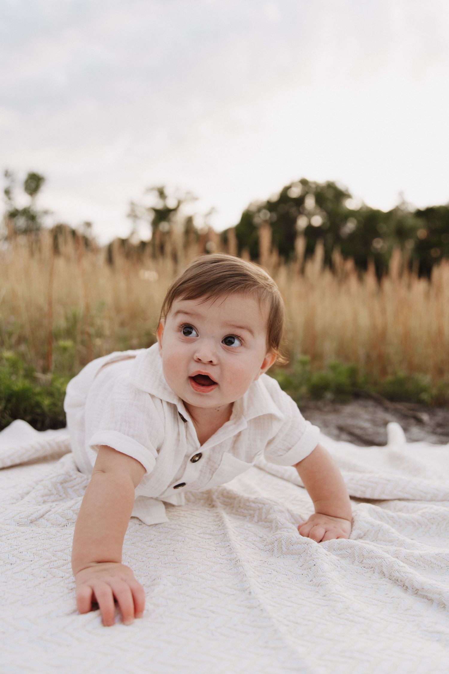 A baby in white clothing crawls on a white blanket outdoors with tall grass in the background.