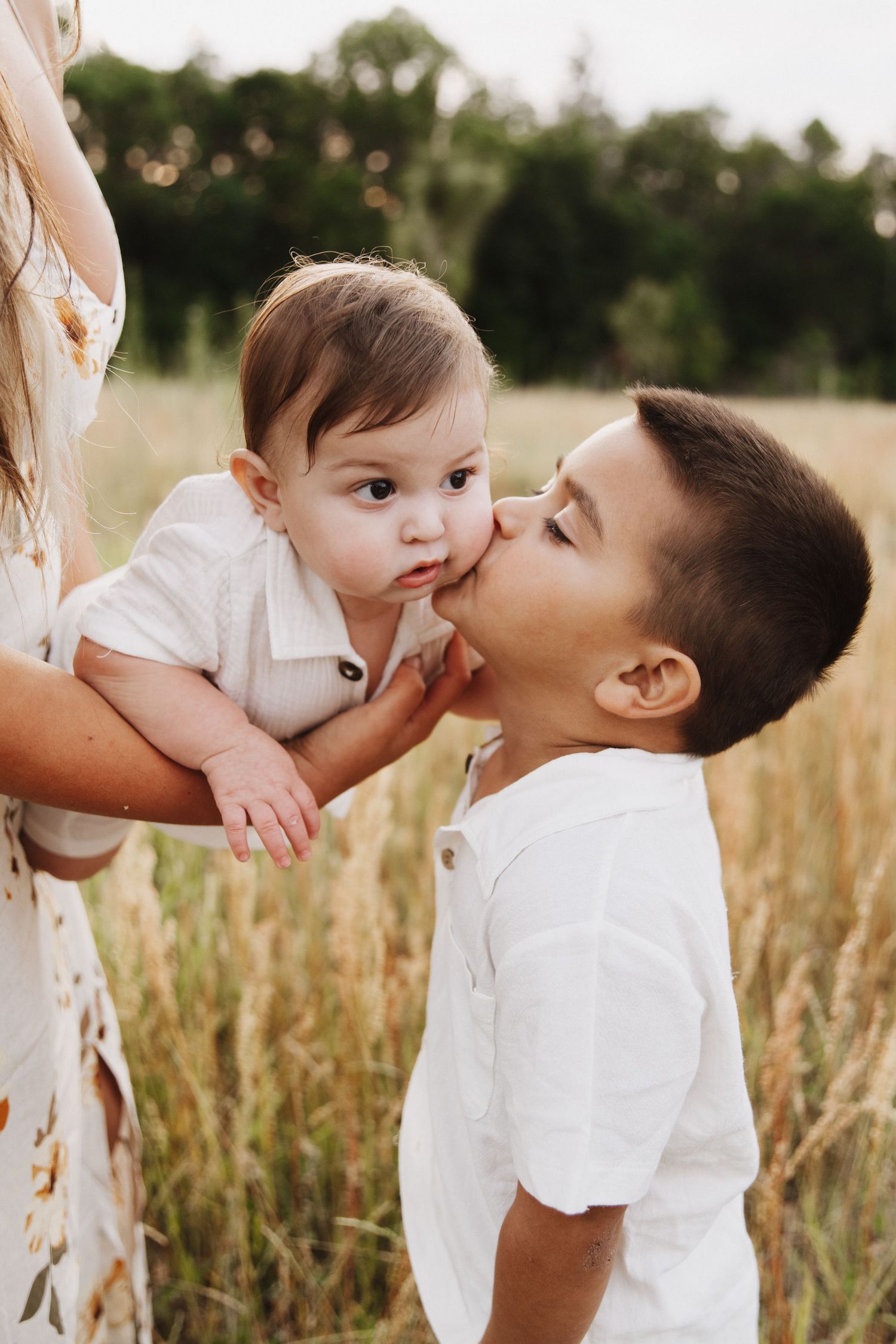 A tender outdoor family moment captured in tall grass with white clothing and soft natural lighting.