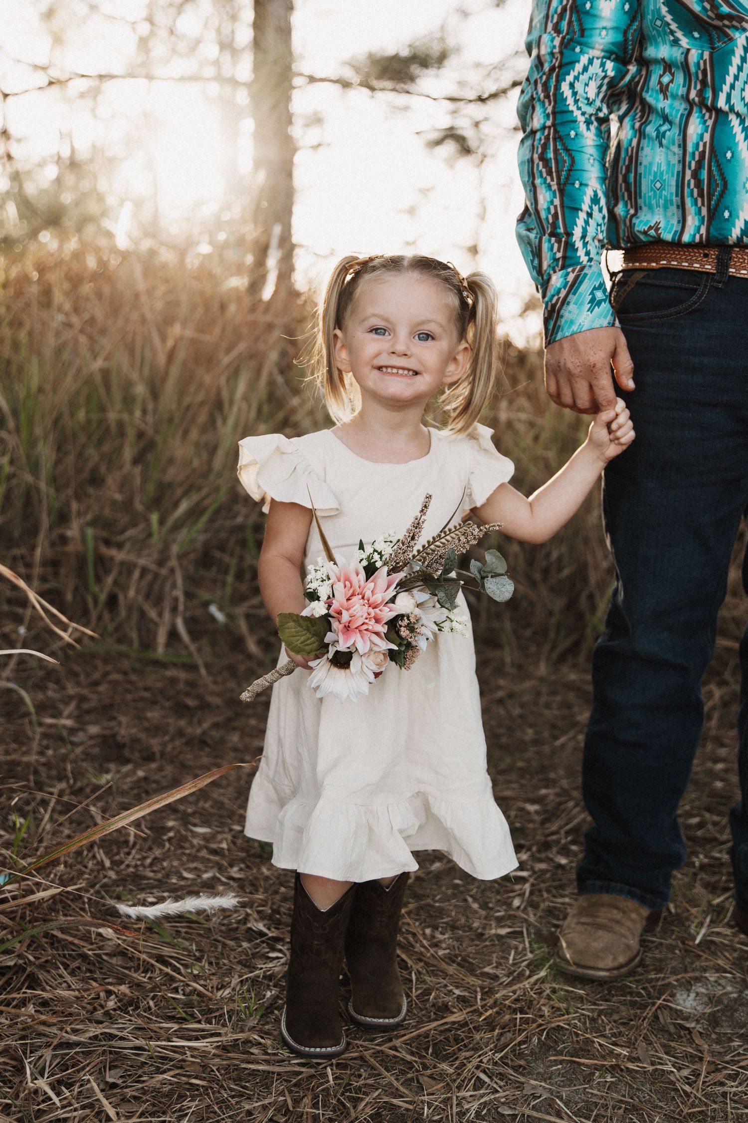 A child's feet and white dress stand next to an adult wearing a turquoise patterned top in a rustic outdoor setting.