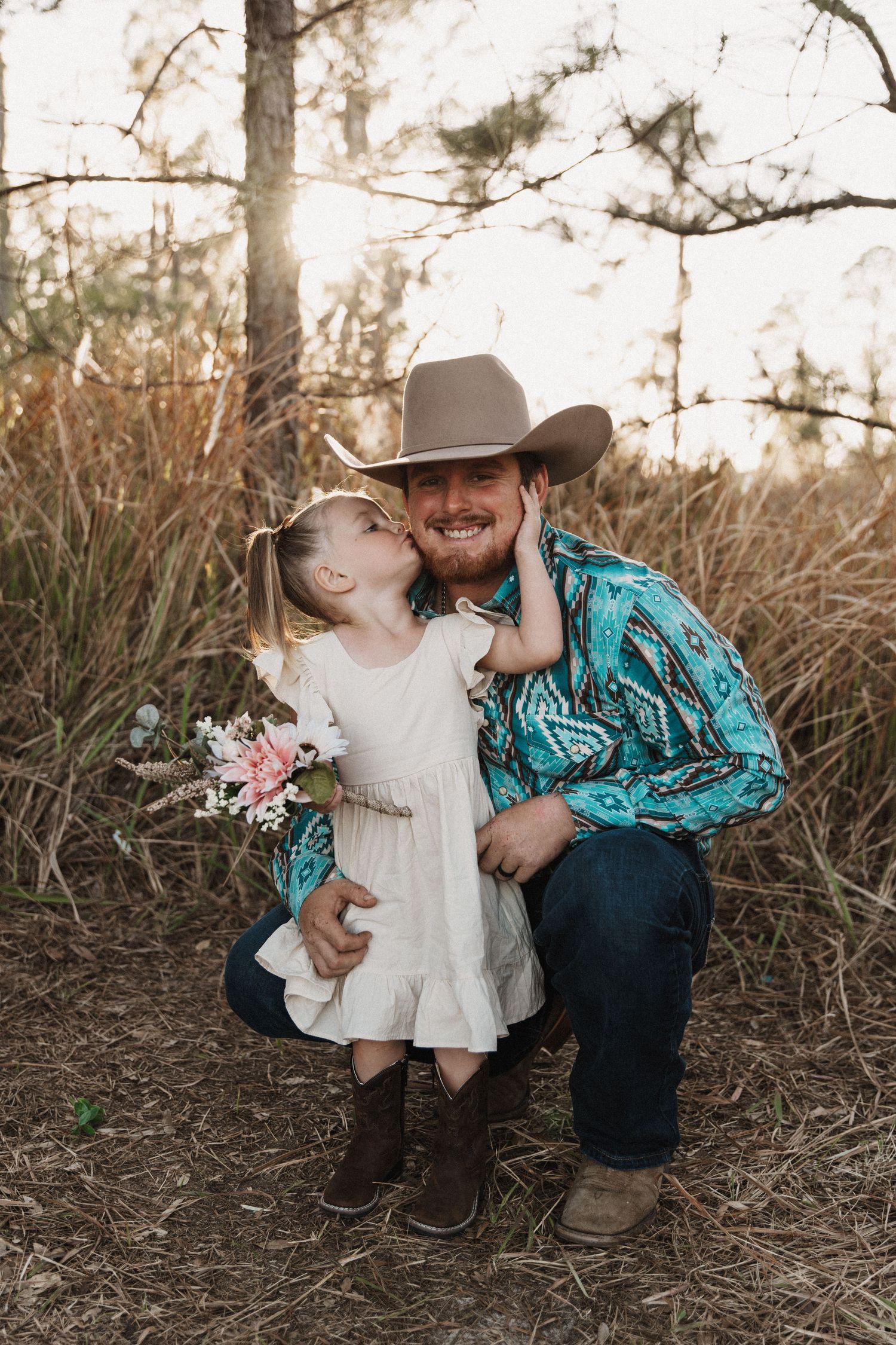 A sweet tender moment shared between a cowboy in turquoise and a young child in white dress against a wooded backdrop.