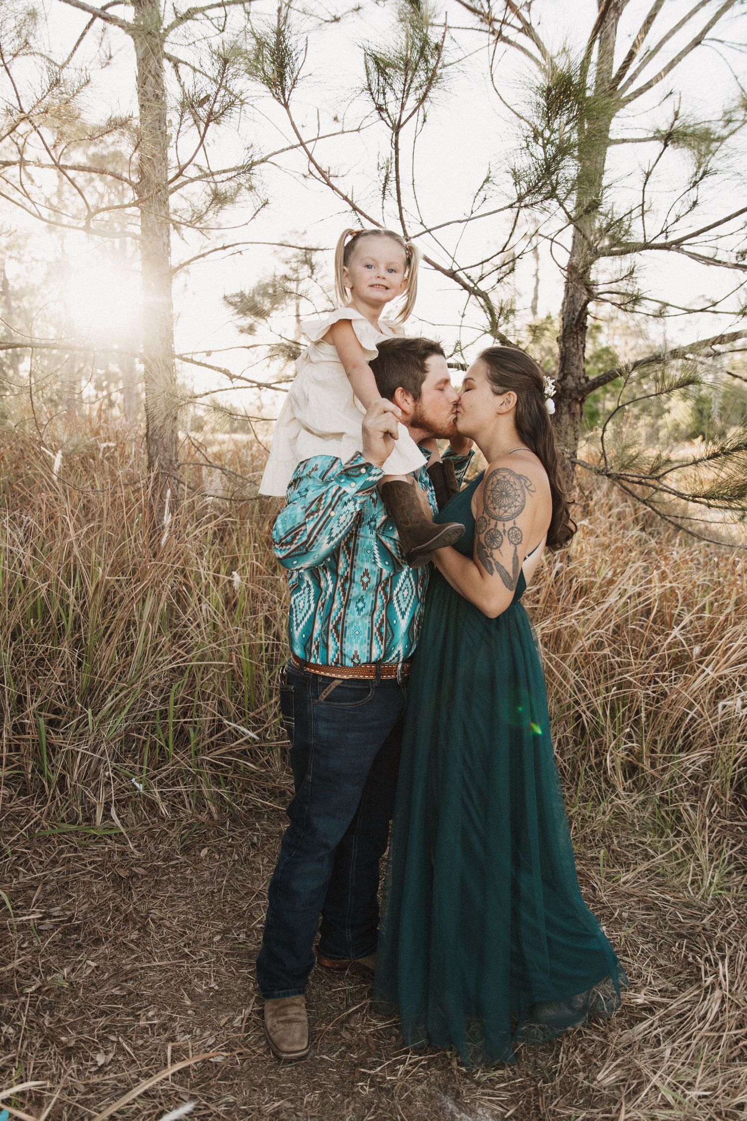 Family of three poses together in a sun-lit forest setting wearing coordinated outfits of emerald green and turquoise.