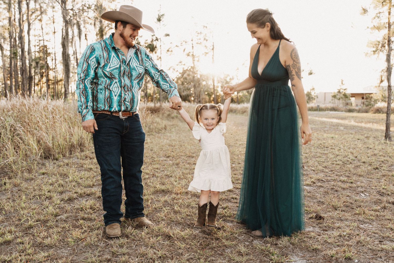 A family in western attire holds hands while standing in a golden field at sunset.