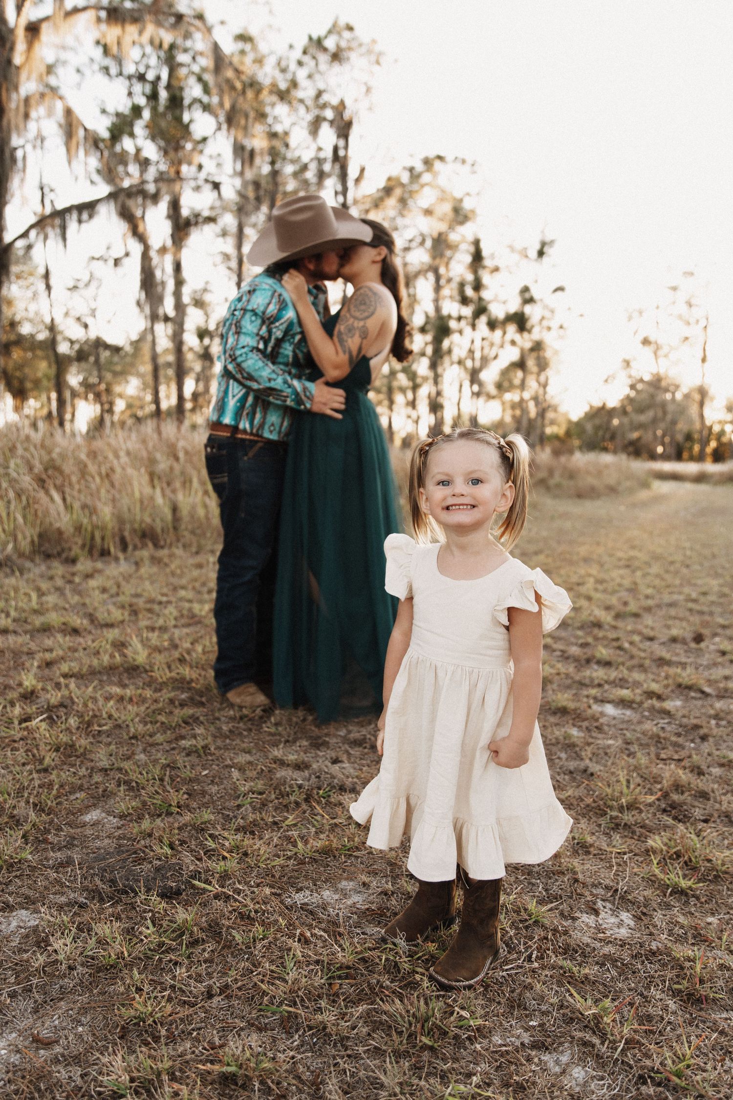 Little girl in white dress stands in grassy field at sunset while country-styled couple embraces in background.