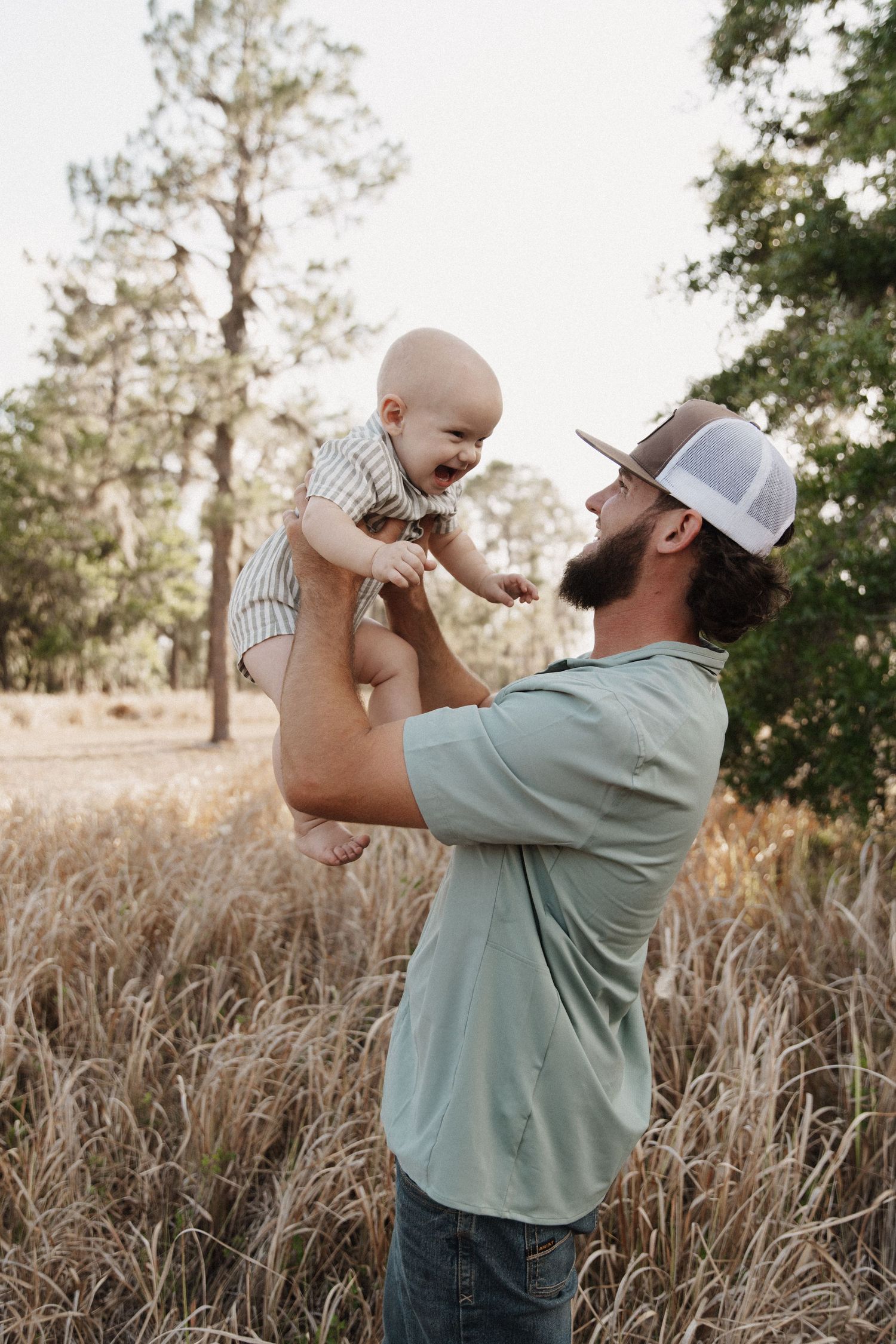 A person in a mint green shirt and baseball cap lifts a small child up in a sequence across a field of tall grass.