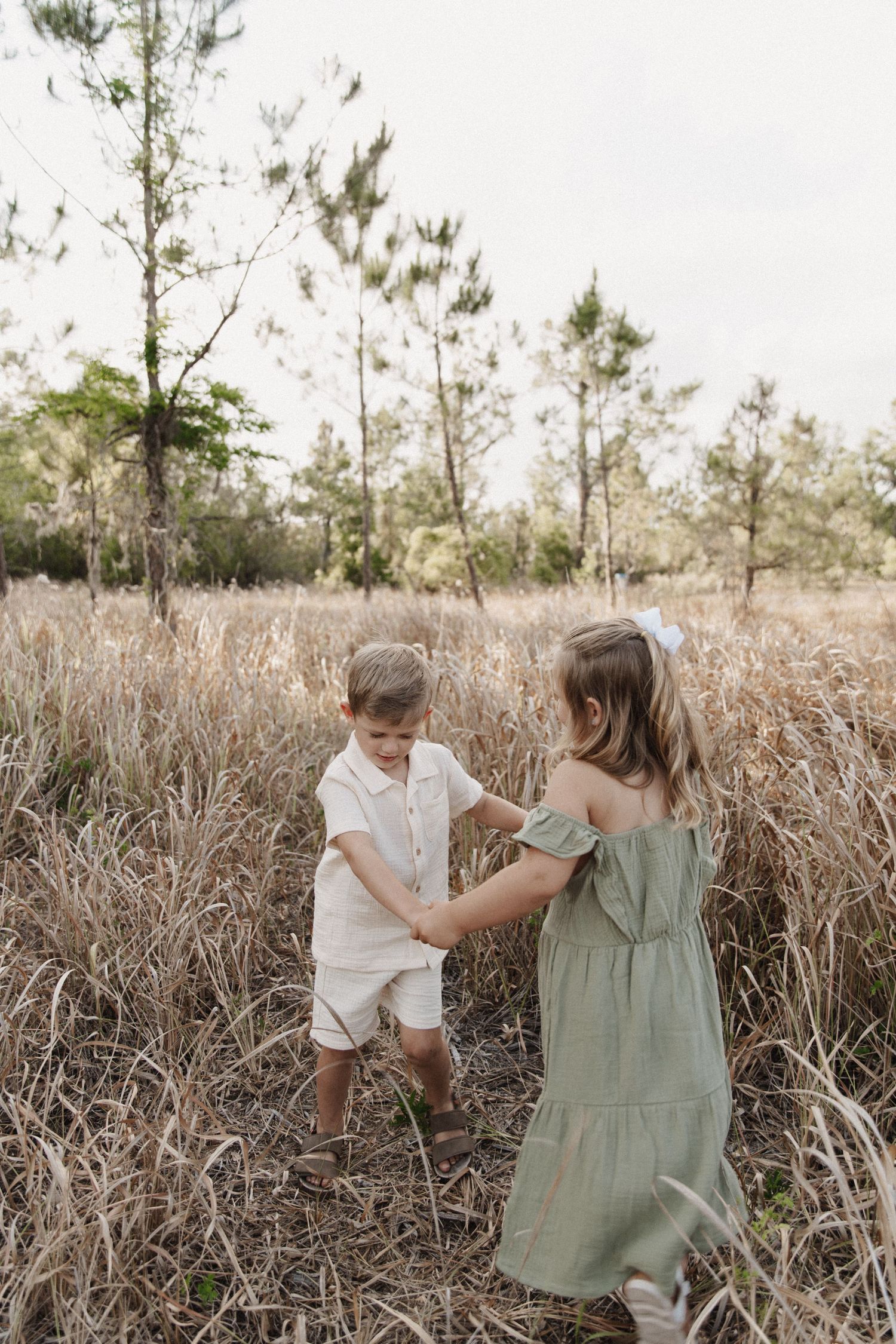 Two children in rustic clothing dance together in a field of tall grass with pine trees in the background.