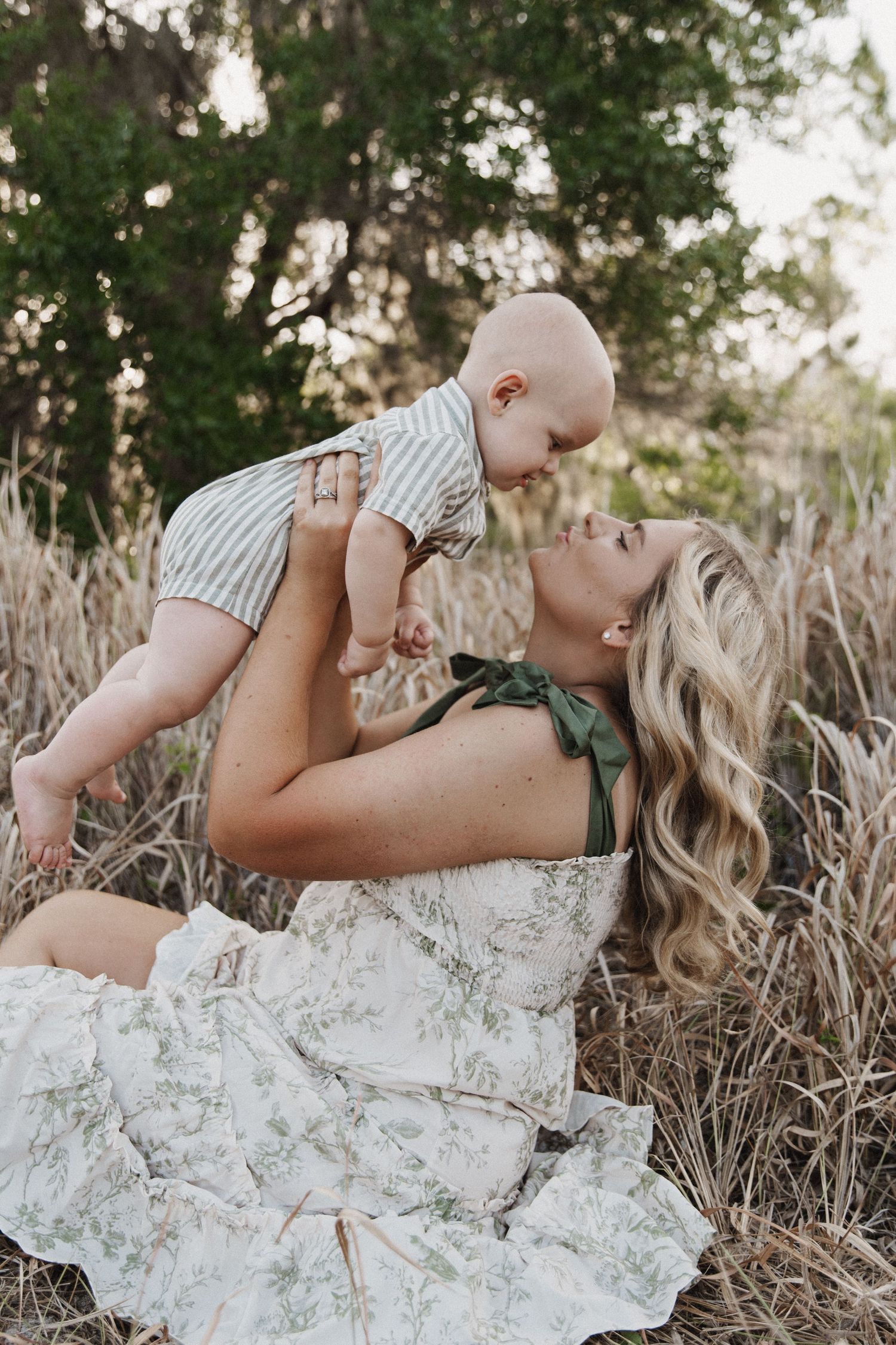 Mother in a white dress lies in tall grass while lifting and playing with baby in striped onesie in outdoor photoshoot.