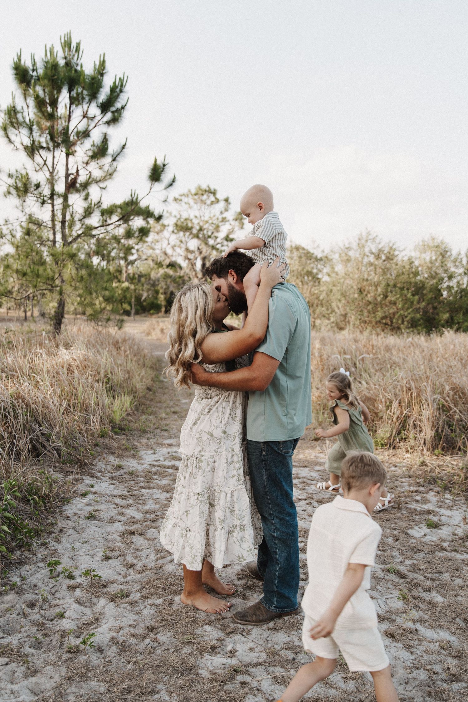 Family sharing tender moment together on outdoor dirt path surrounded by tall grass and pine trees at sunset.
