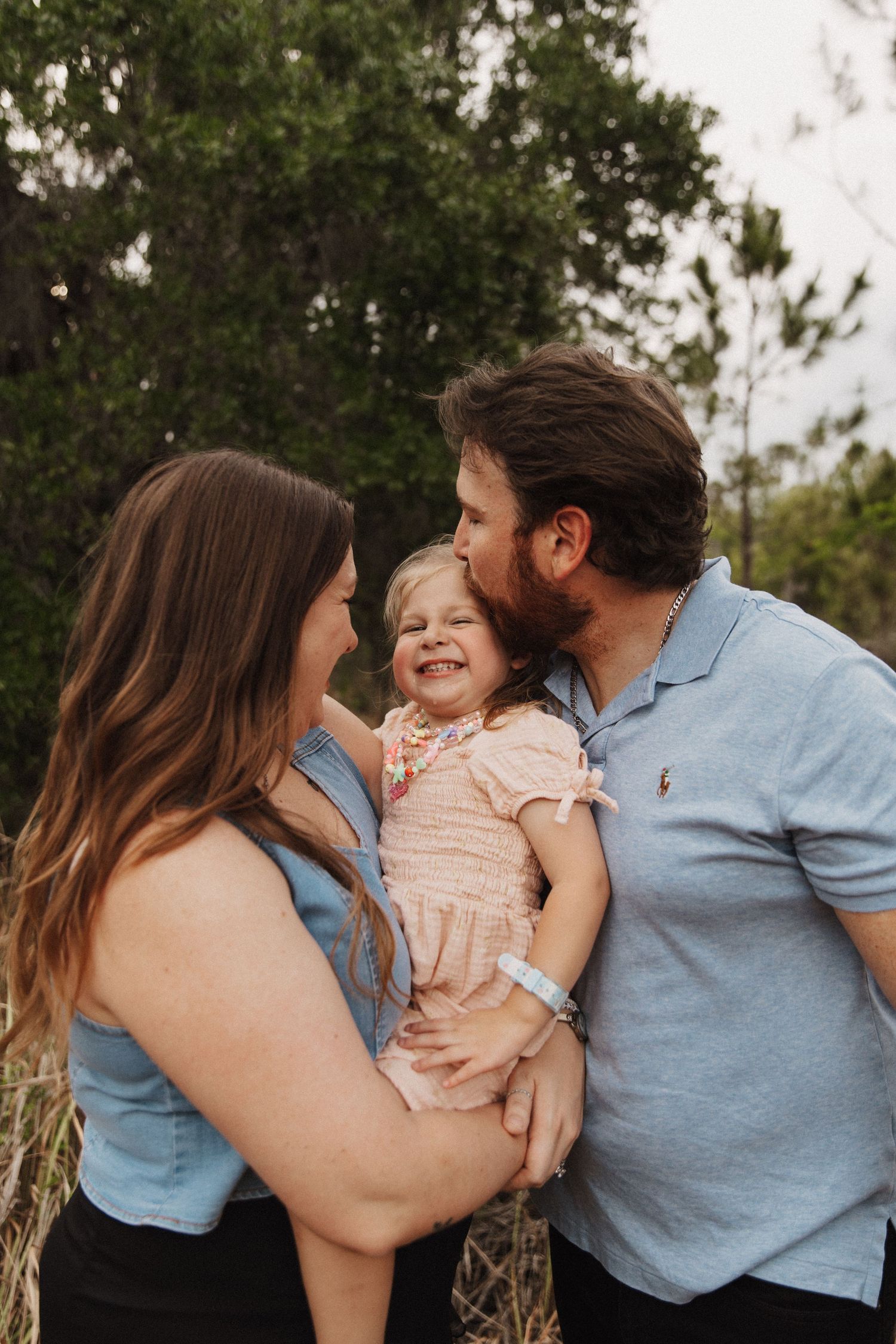 Family sharing playful kisses and hugs with young child in casual outdoor photo session.