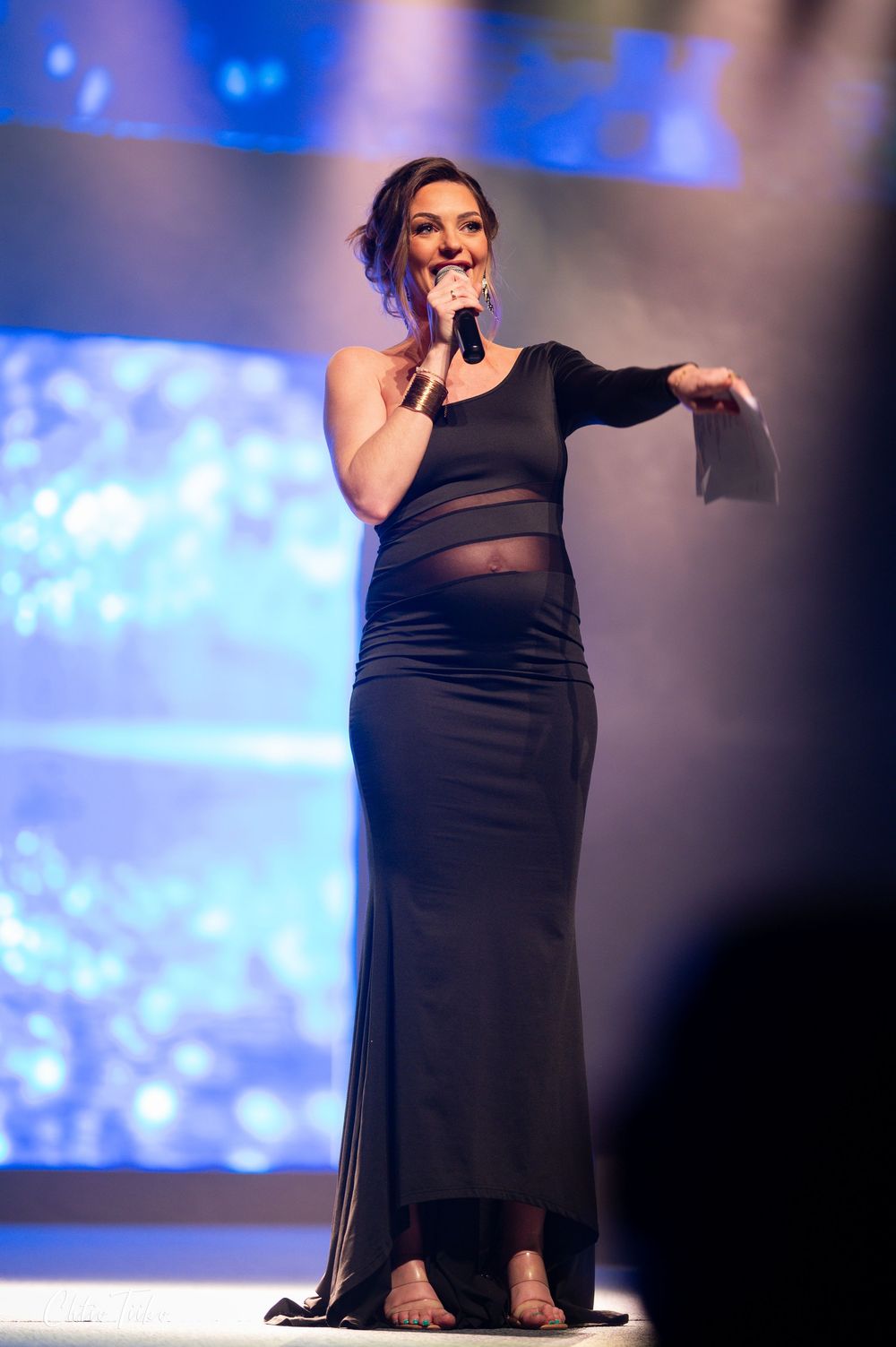 Woman performs on stage in elegant black one-shoulder gown with sheer panel while holding microphone.