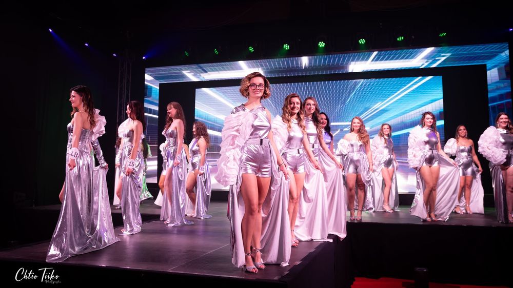 Group of pageant contestants walk runway together in silver and white gowns at beauty competition.