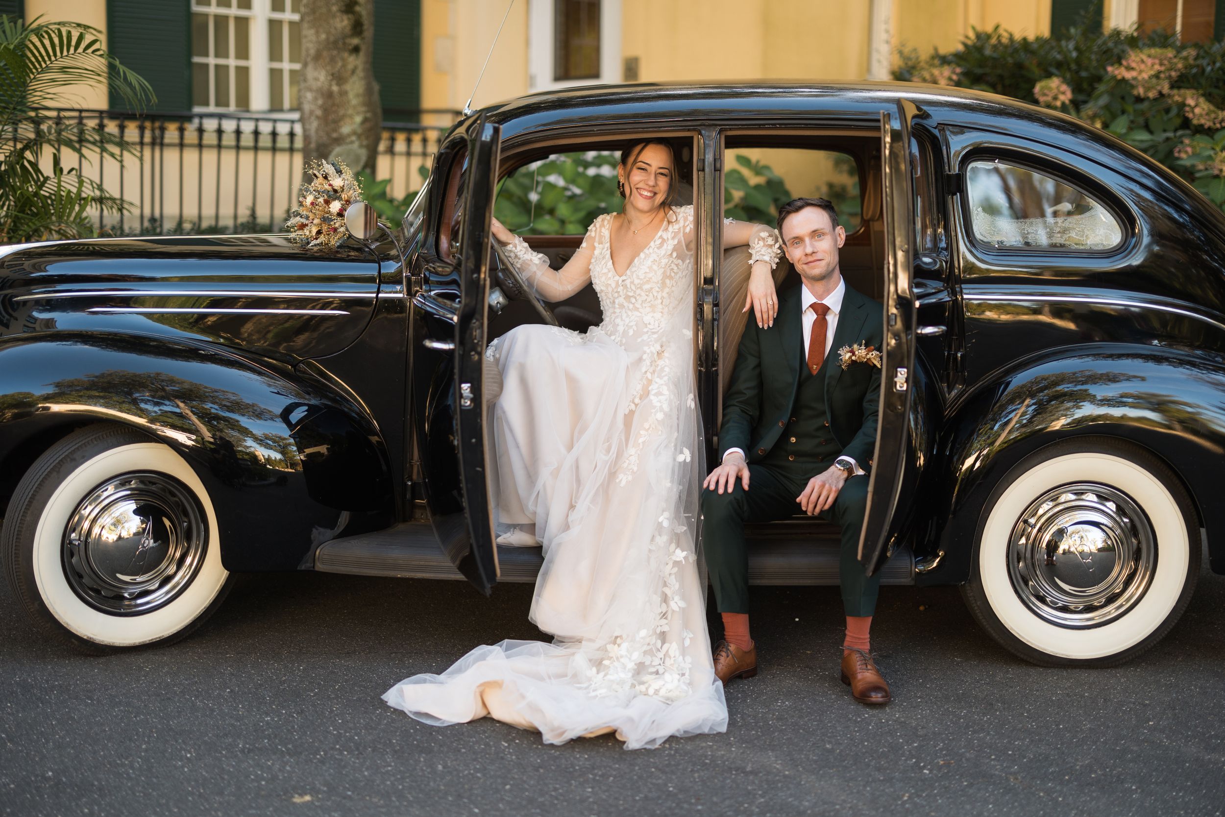 Elegant couple poses with classic black vintage car on their wedding day.