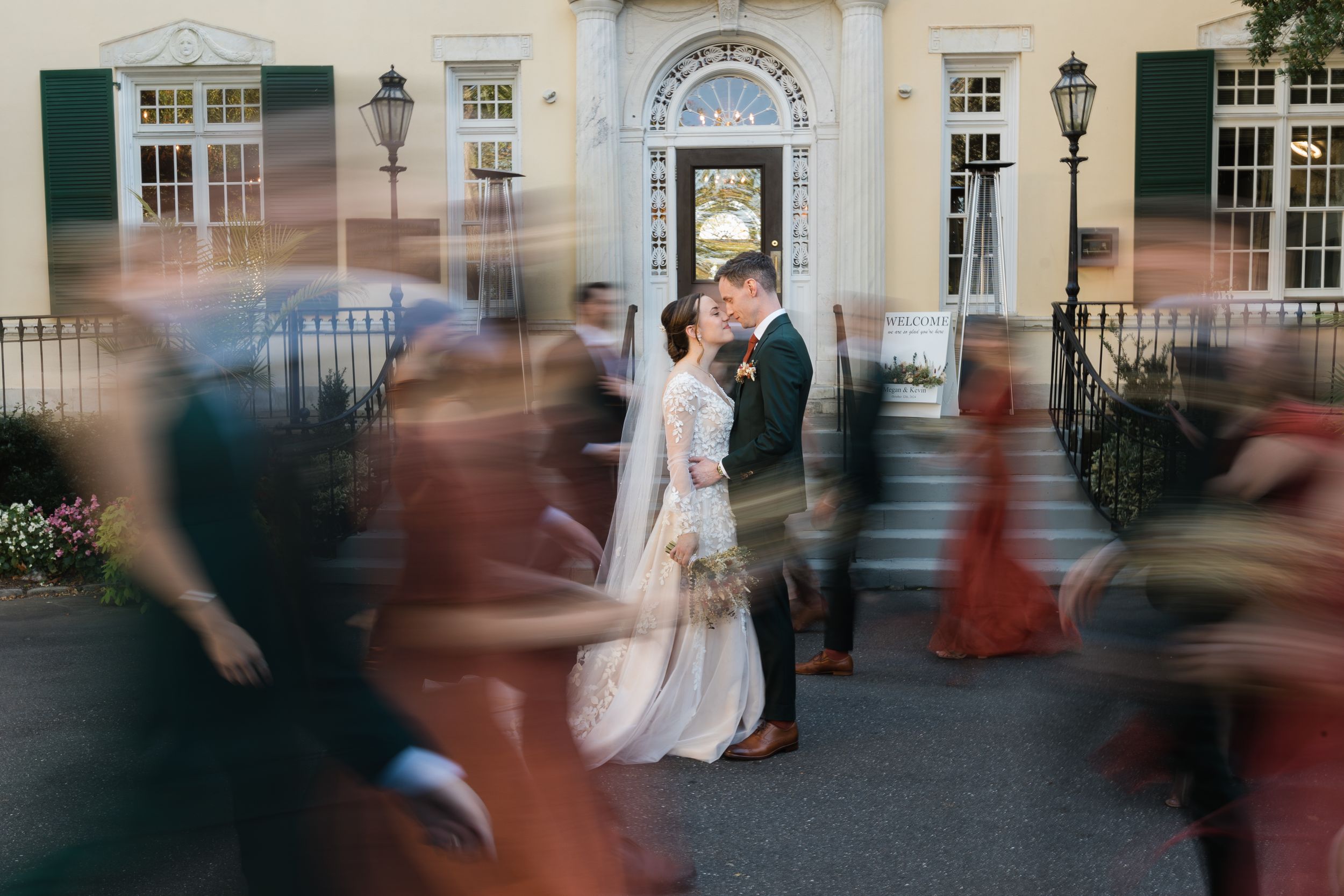 Wedding couple shares moment amid motion-blurred guests outside historic building.