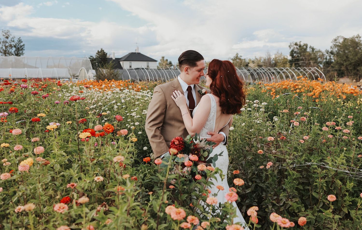 Romantic couple embraces in a vibrant orange zinnia field in Colorado Springs on a sunny day with mountains in the background.