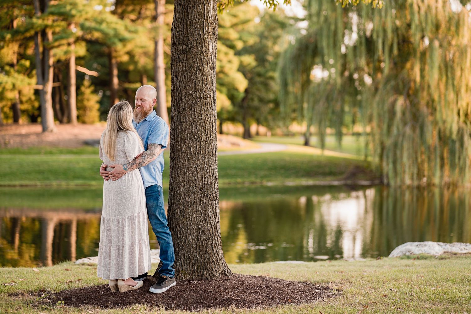 A couple shares an intimate moment beside a serene lake with weeping willow trees in the background.