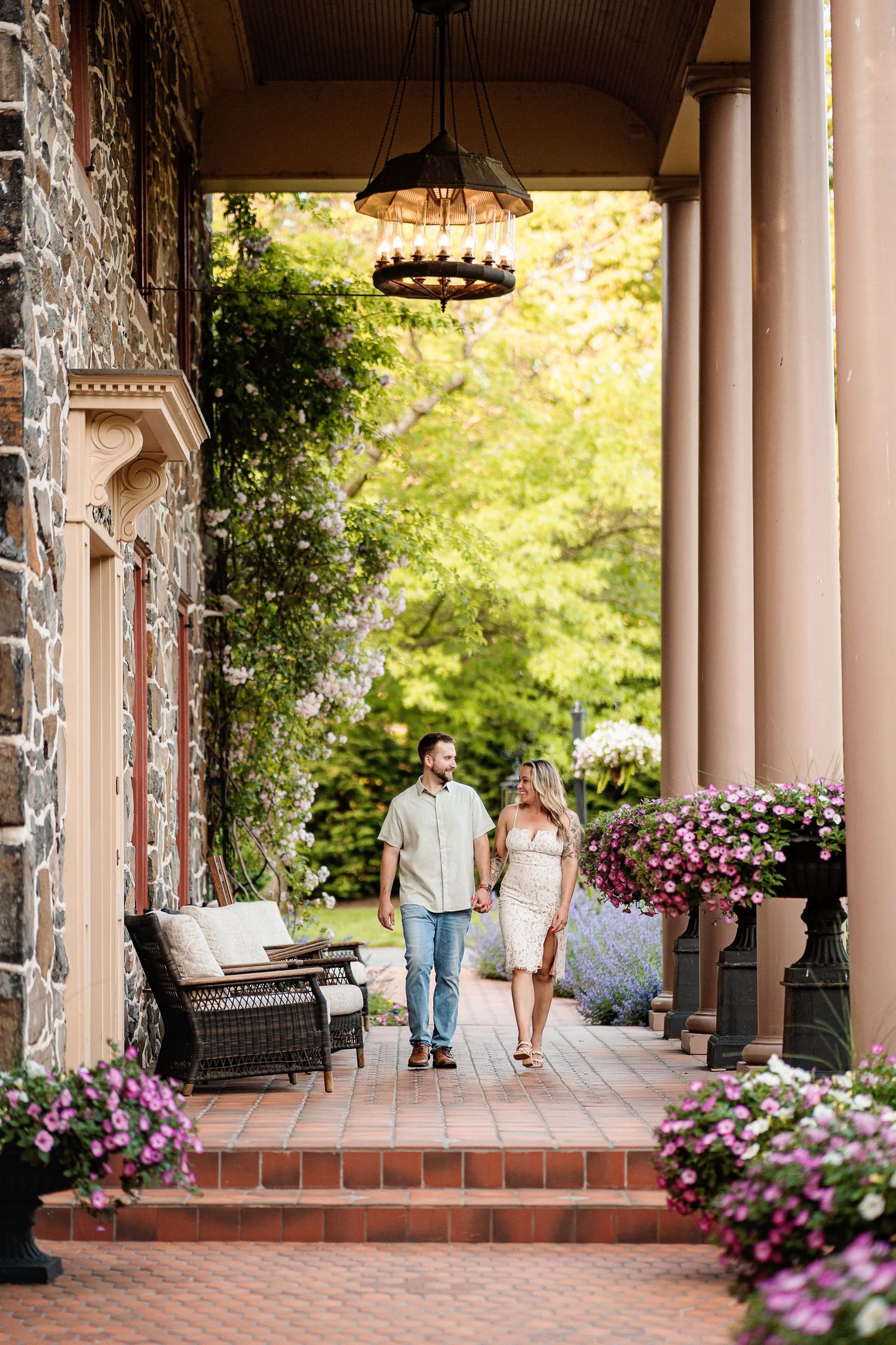 A romantic couple walks through a columned veranda adorned with hanging purple flowers at an elegant estate.