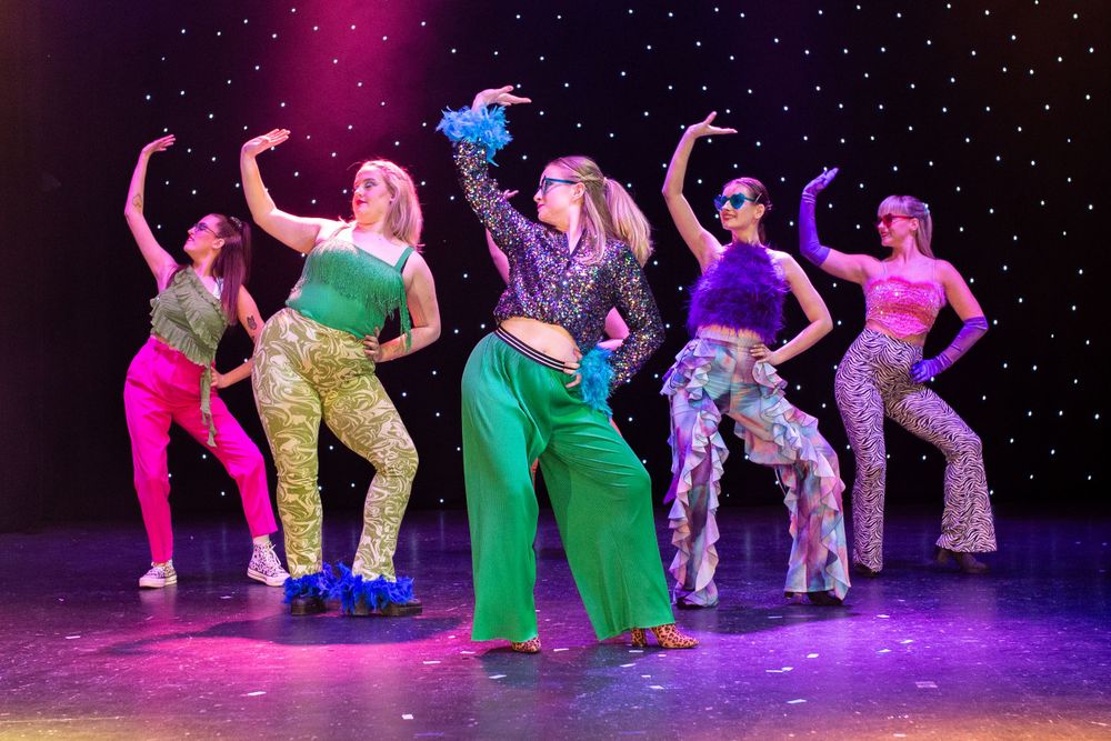 Dancers perform on stage in vibrant costumes under colorful theatrical lighting.