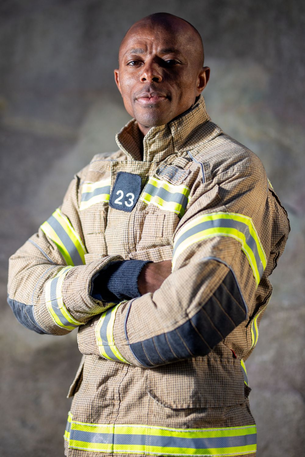 Firefighter poses in protective gear with reflective stripes against grey background.