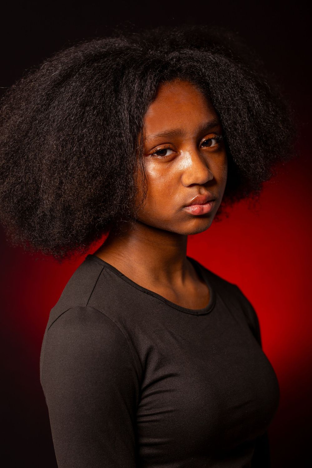 Young Black woman with voluminous afro hair wearing black top, posing against dramatic red background.
