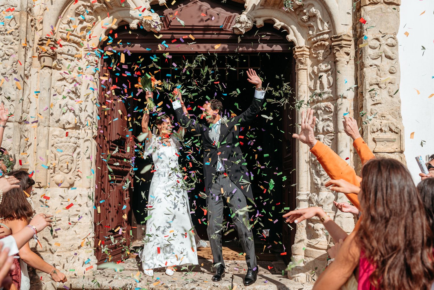 Celebratory scene at ornate church doorway with confetti being thrown as couple exits after wedding ceremony.