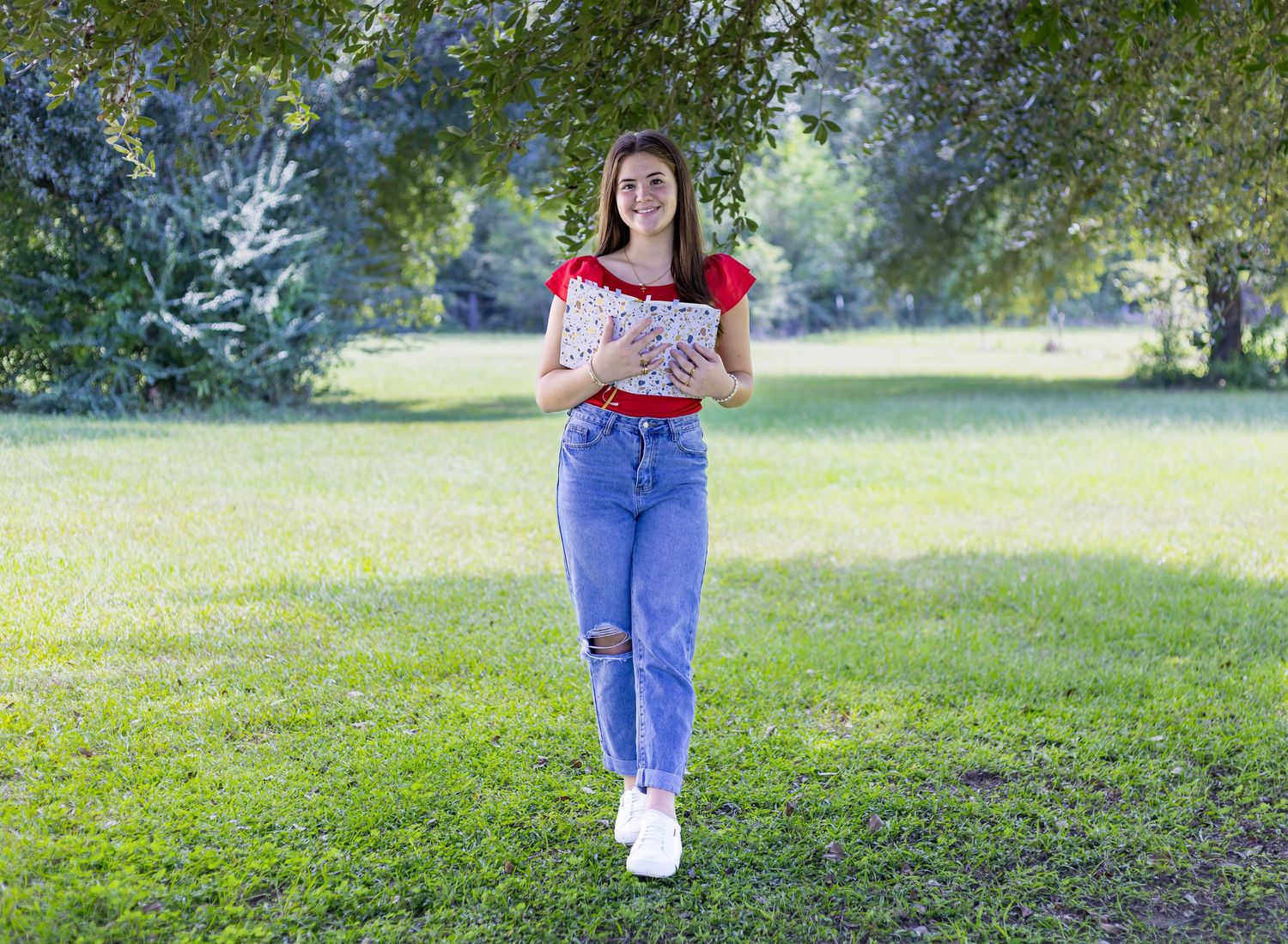 A person in ripped jeans and white sneakers walks through a sunny green park on a summer day.