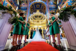 Elegant church wedding ceremony with bridesmaids in emerald green dresses lining a red-carpeted aisle under ornate chandeliers.