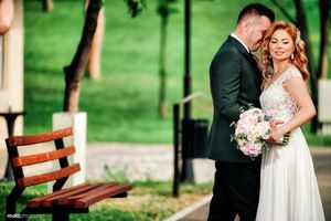 A romantic wedding couple embraces outdoors in a park setting with a wooden bench and pink bouquet.