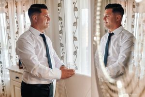 A man in a white dress shirt and tie stands looking at his reflection in a mirror with sheer curtains behind him.