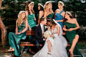 Wedding party poses playfully on wooden bench with bridesmaids in emerald green dresses and white gown.